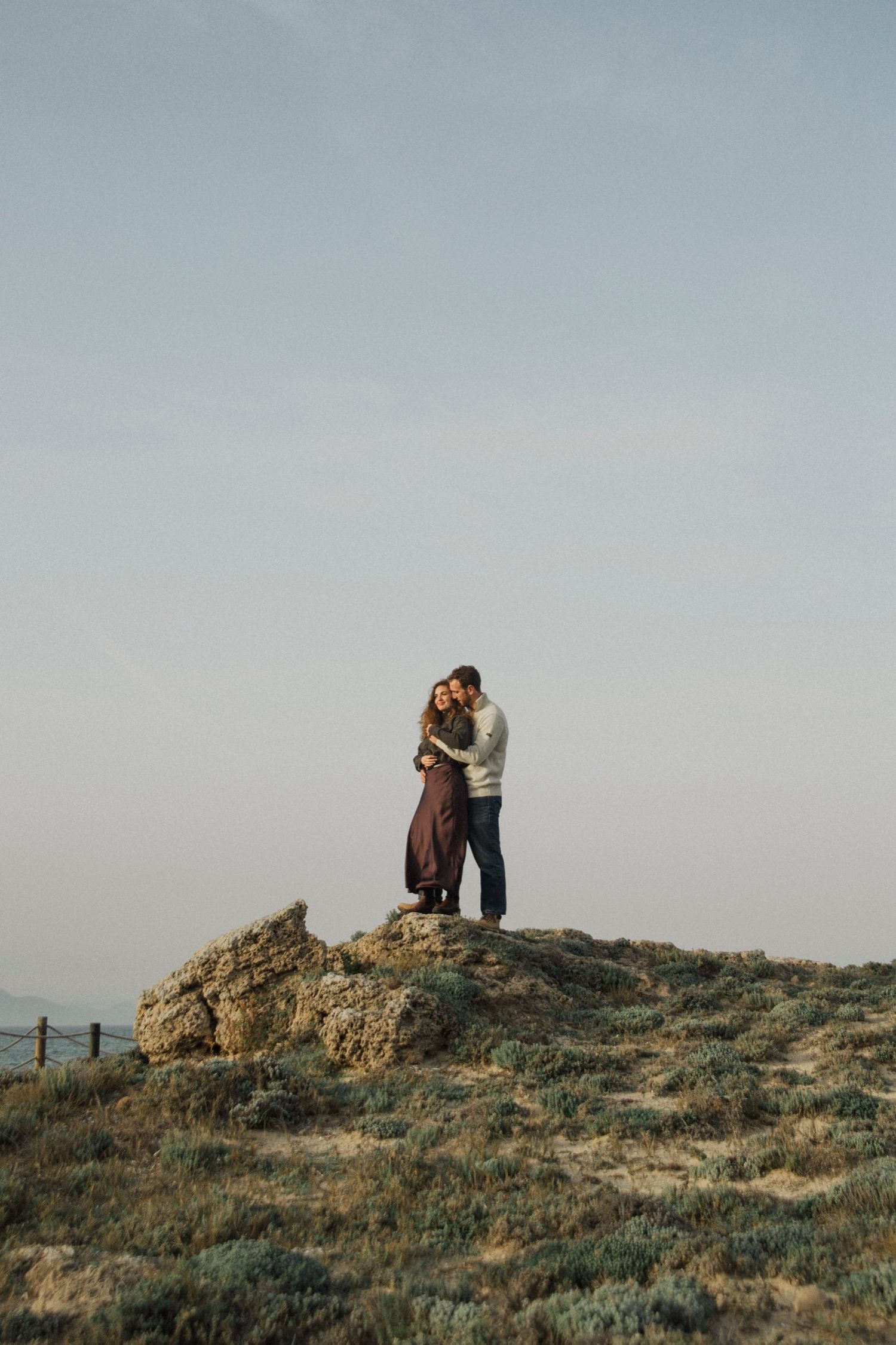 Two people share an intimate moment together on a hilltop overlooking a scenic coastal landscape at sunset.