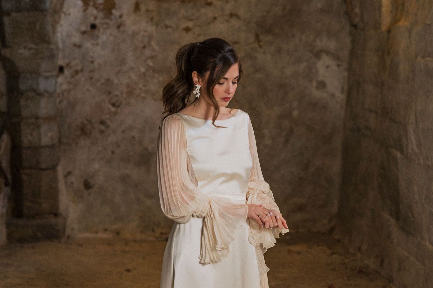 A bride in a white dress and veil stands against a rustic stone wall in a romantic wedding setting.