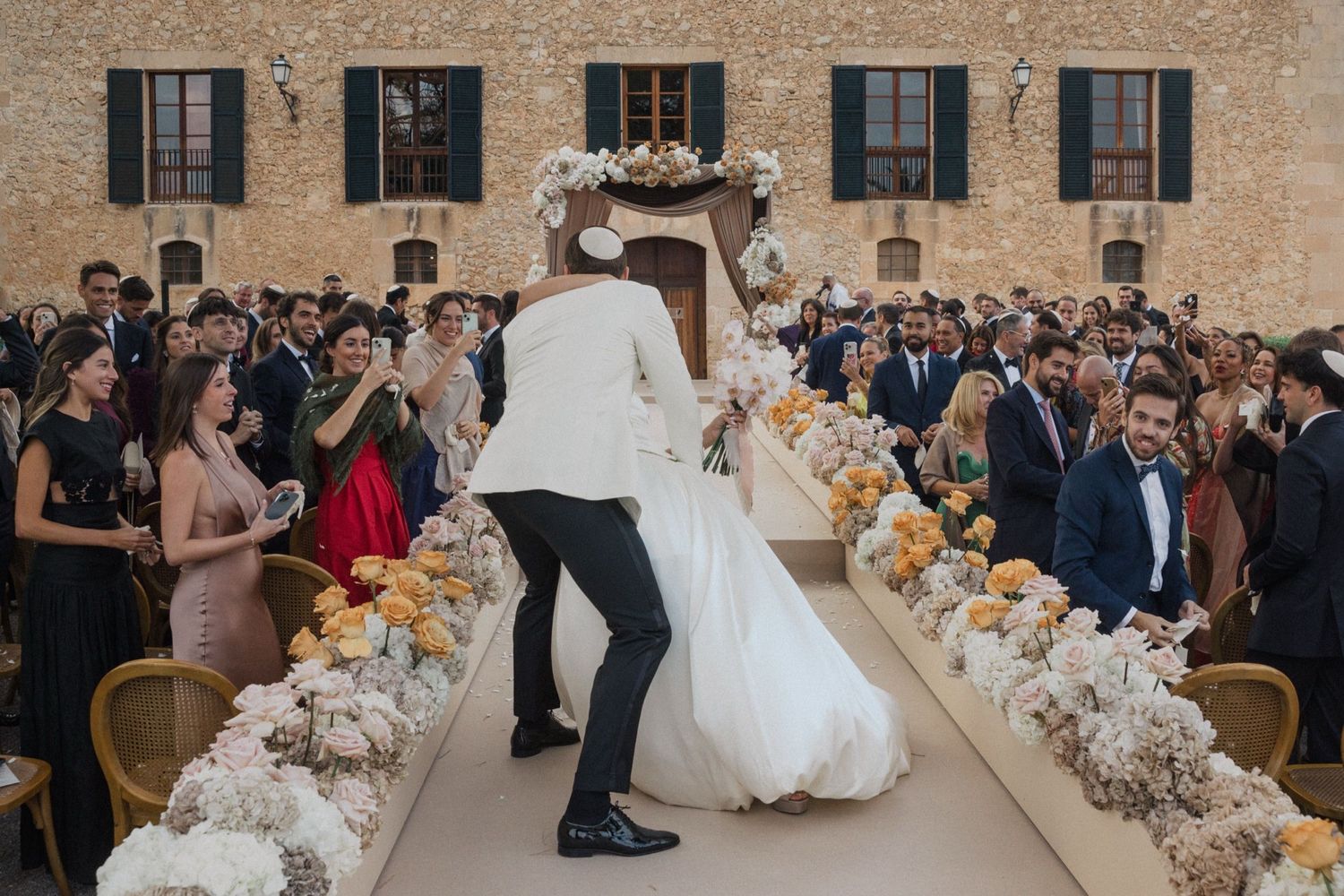 Elegant outdoor wedding ceremony with white floral aisle decorations and guests gathered at a stone venue.