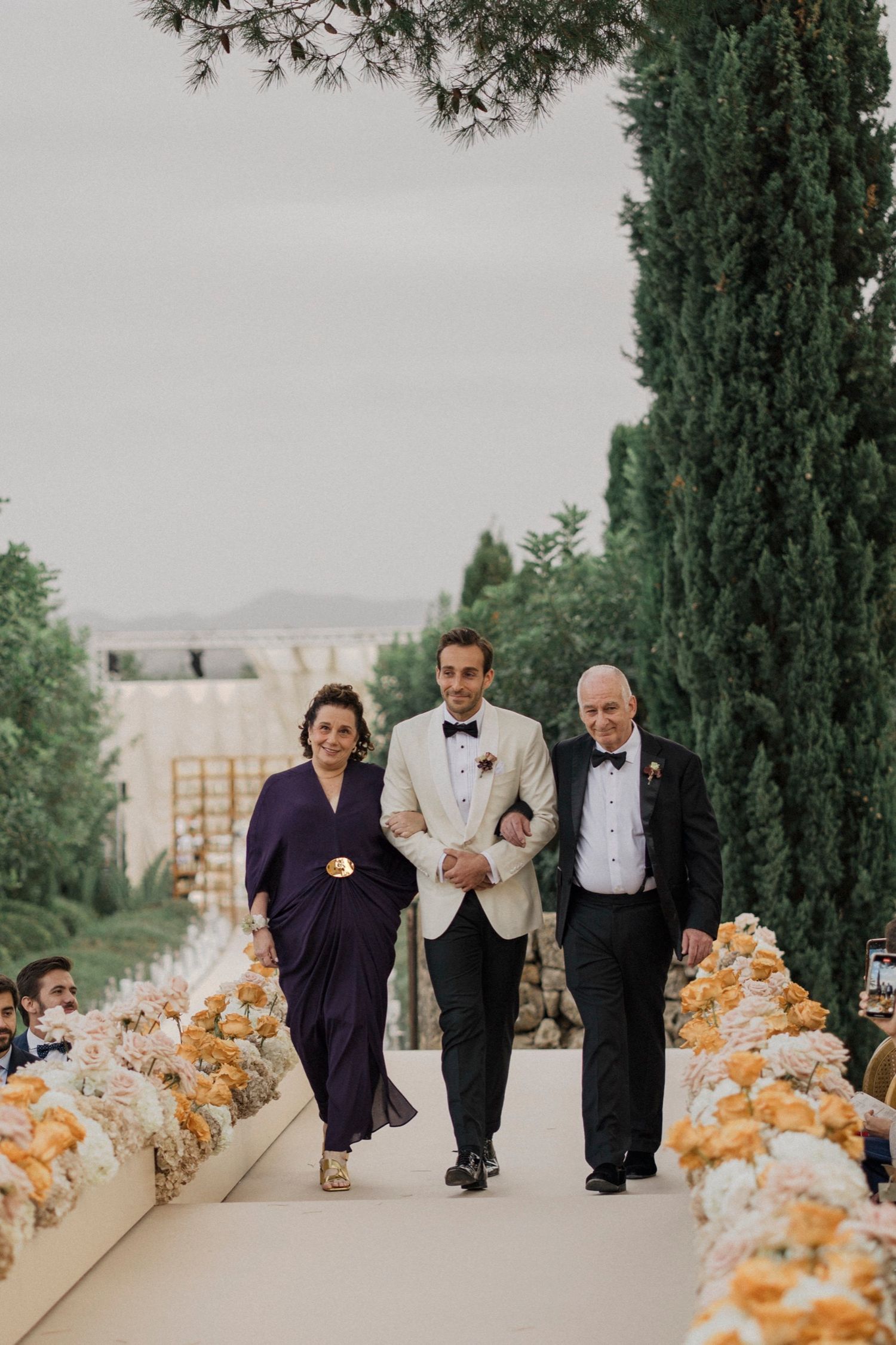 Wedding party members walk down an aisle lined with peach and orange floral arrangements at an outdoor ceremony.