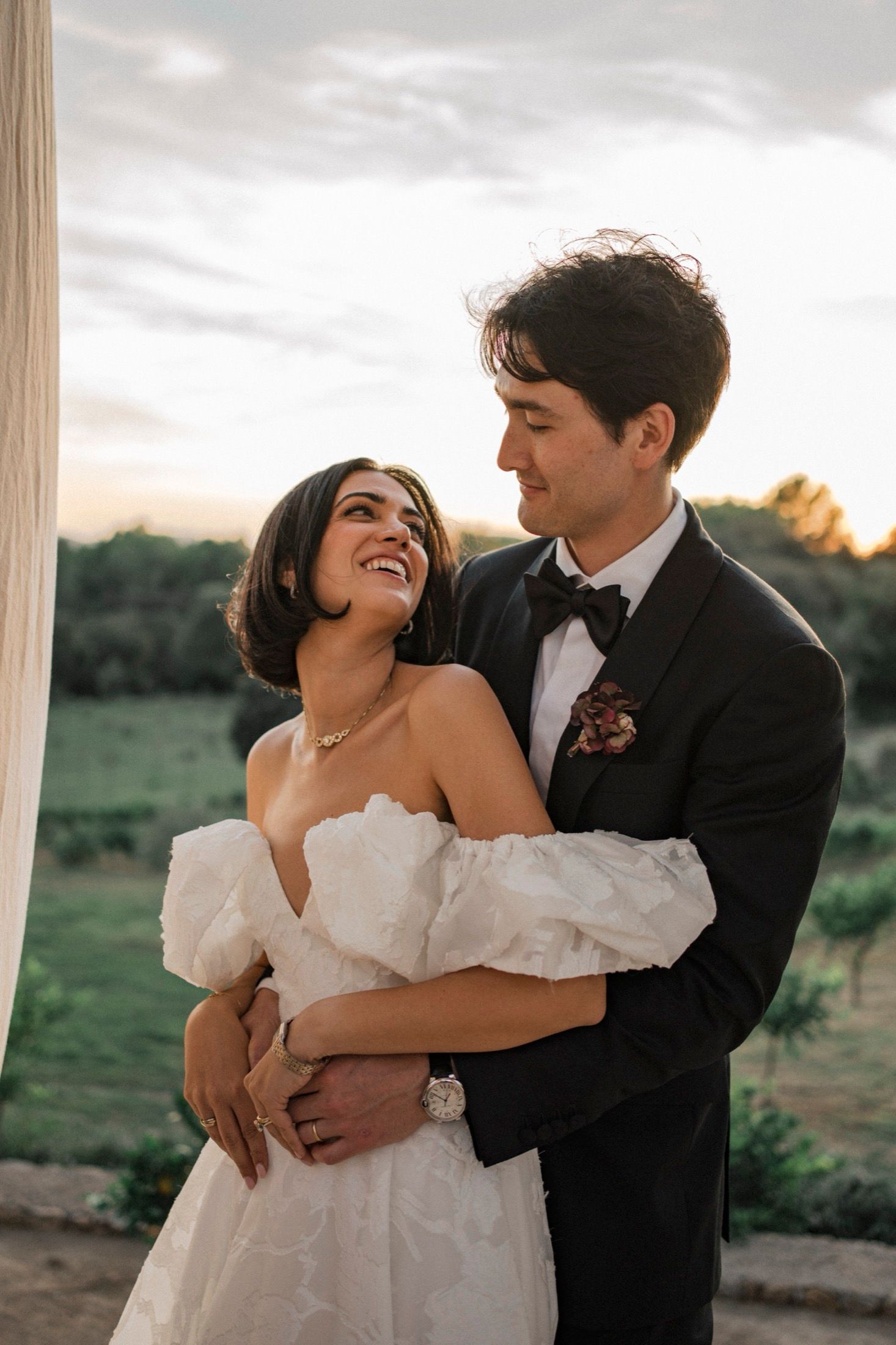 A romantic sunset wedding portrait shows a couple embracing outdoors, with the bride in an off-shoulder white gown.