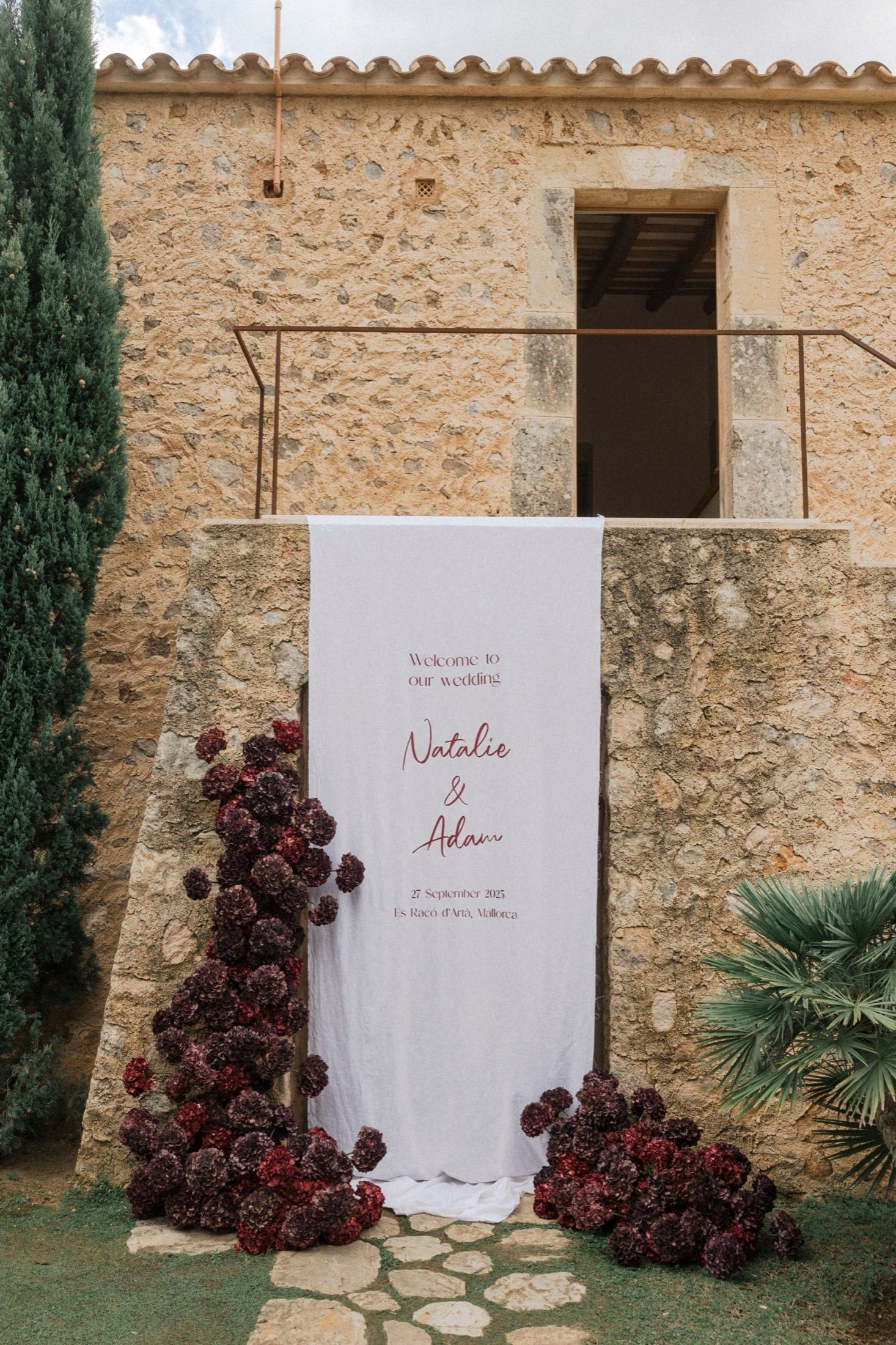 White wedding backdrop with burgundy floral arrangements against a rustic stone wall creates an elegant ceremony setting.