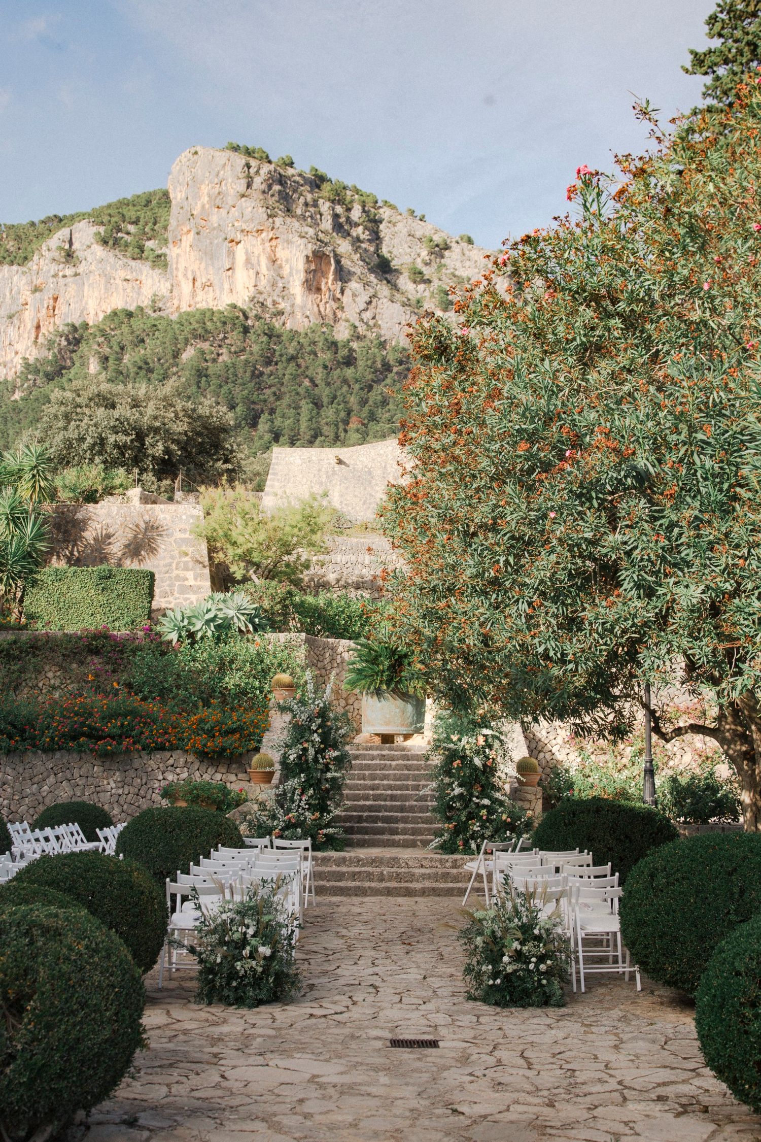 Outdoor wedding ceremony setup with white chairs and stone steps surrounded by flowering trees and mountain backdrop.