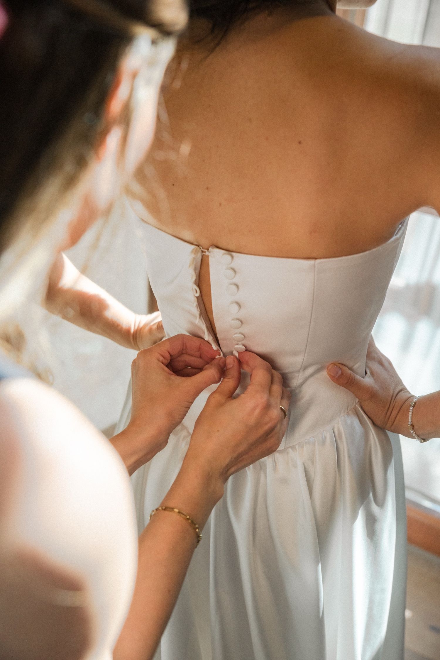 Close-up shot of hands fastening buttons on the back of an elegant white wedding dress.