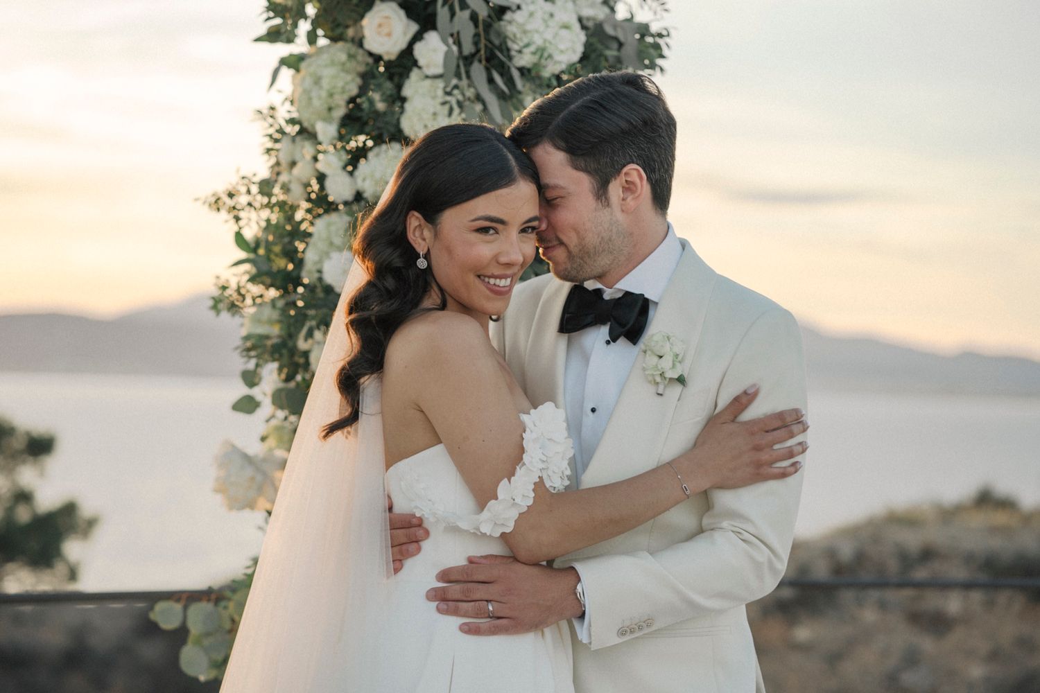 Couple embraces at outdoor wedding ceremony with white floral arch and ocean sunset backdrop.