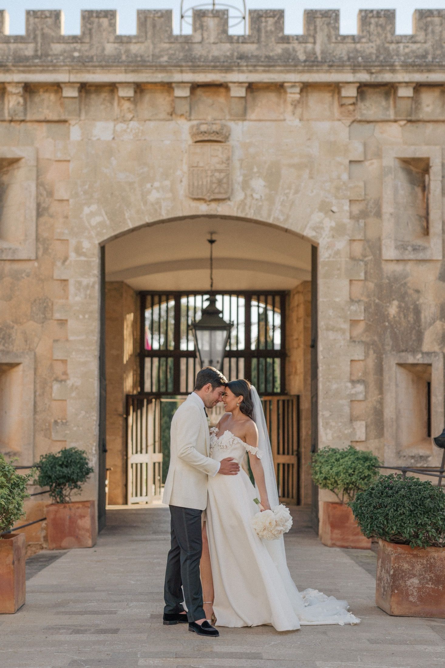 Bride and groom share a romantic embrace in front of a grand stone archway at an elegant wedding venue.