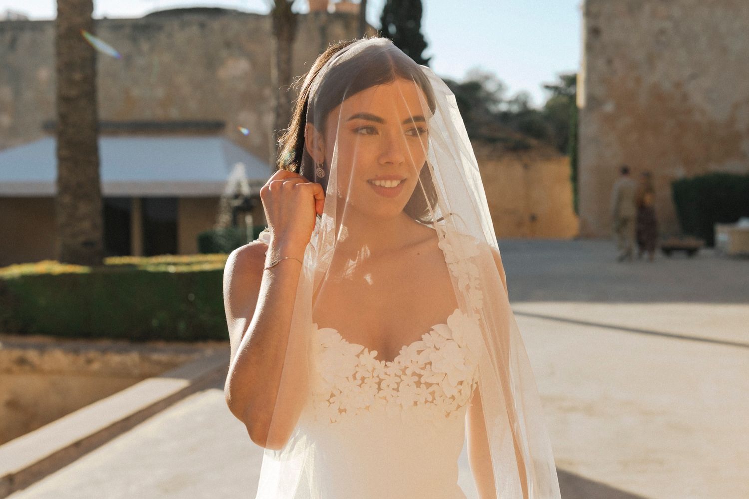 Bride in white lace wedding dress and veil smiling outdoors in sunny courtyard.