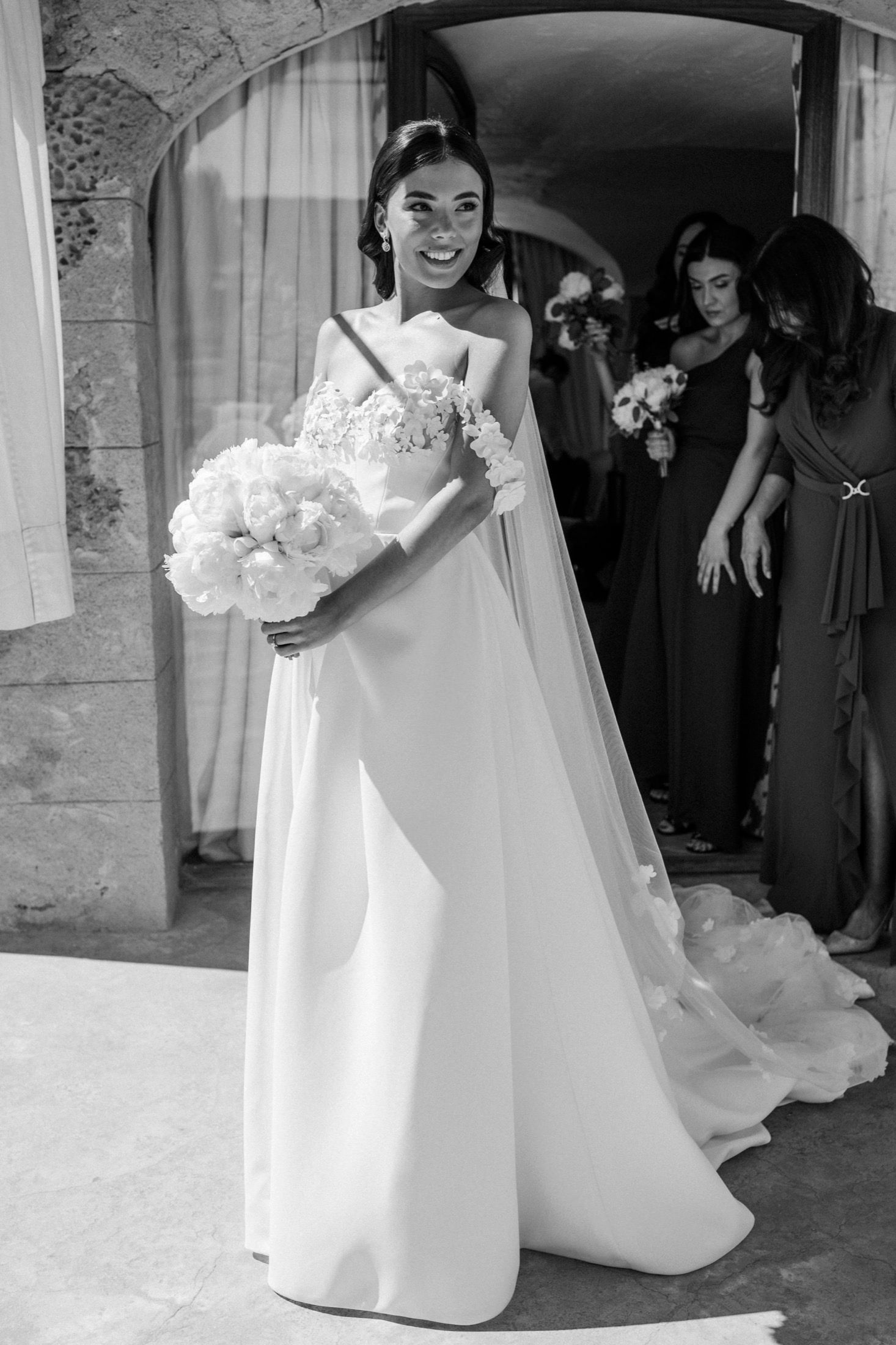 Smiling bride in elegant white gown with floral bodice holds bouquet, bridesmaids visible in doorway behind her.
