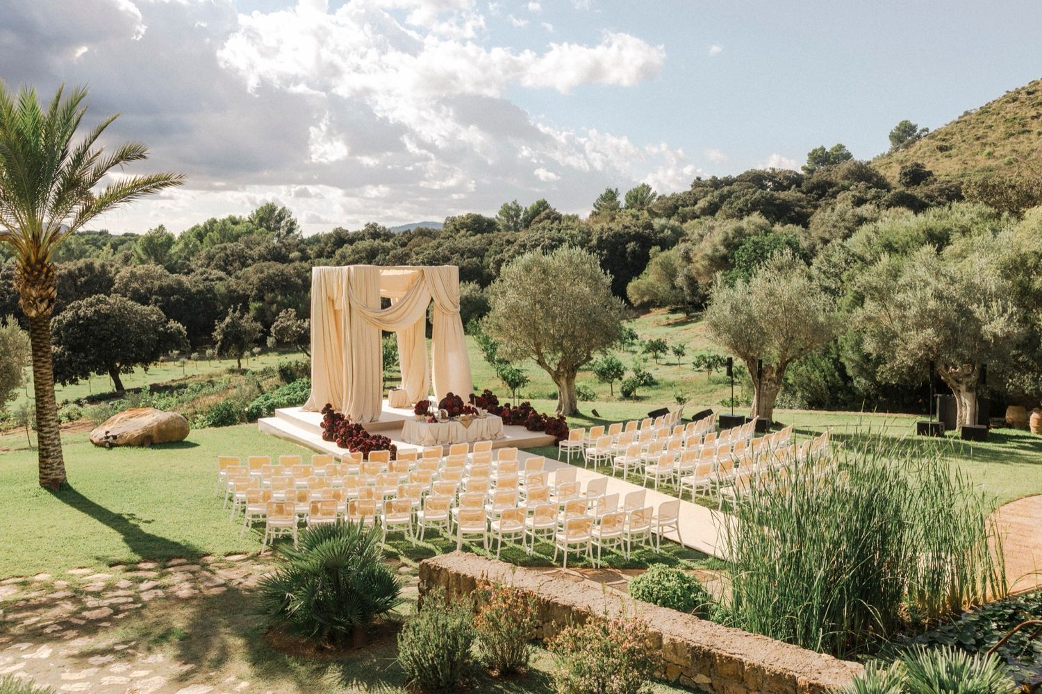 Outdoor wedding ceremony setup with wooden arch and white chairs on green lawn surrounded by olive trees and palm trees.