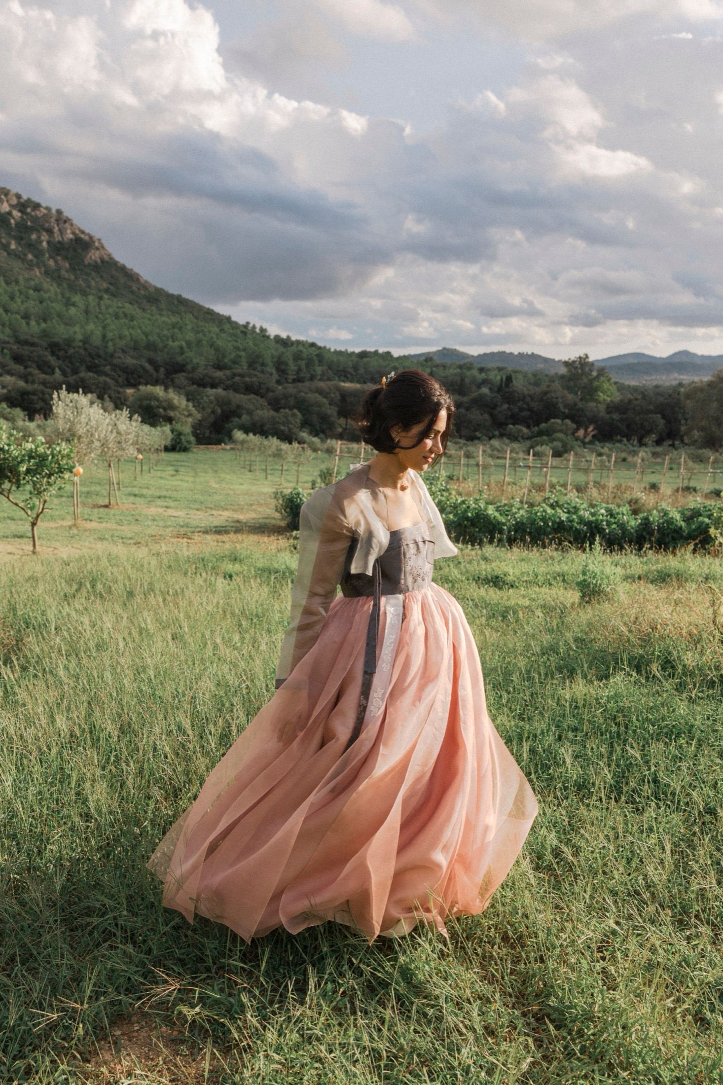 A woman in a flowing pink vintage-style dress twirls gracefully in a mountain meadow at sunset.