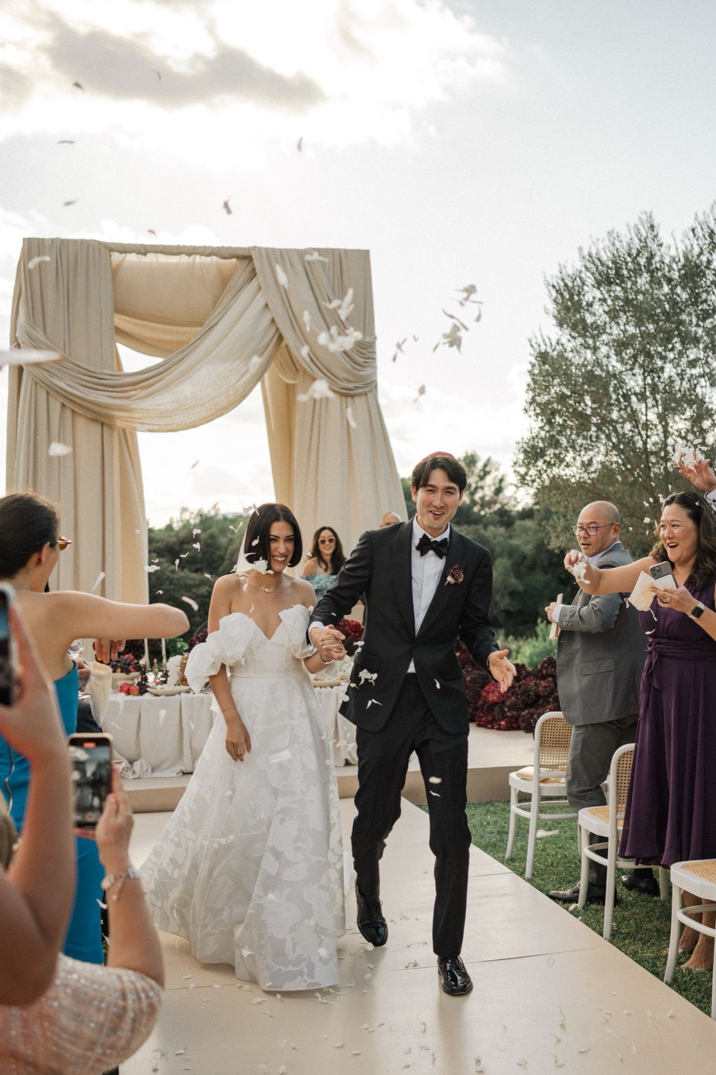 Wedding couple exits their ceremony under a draped white arch while guests toss petals in celebration.