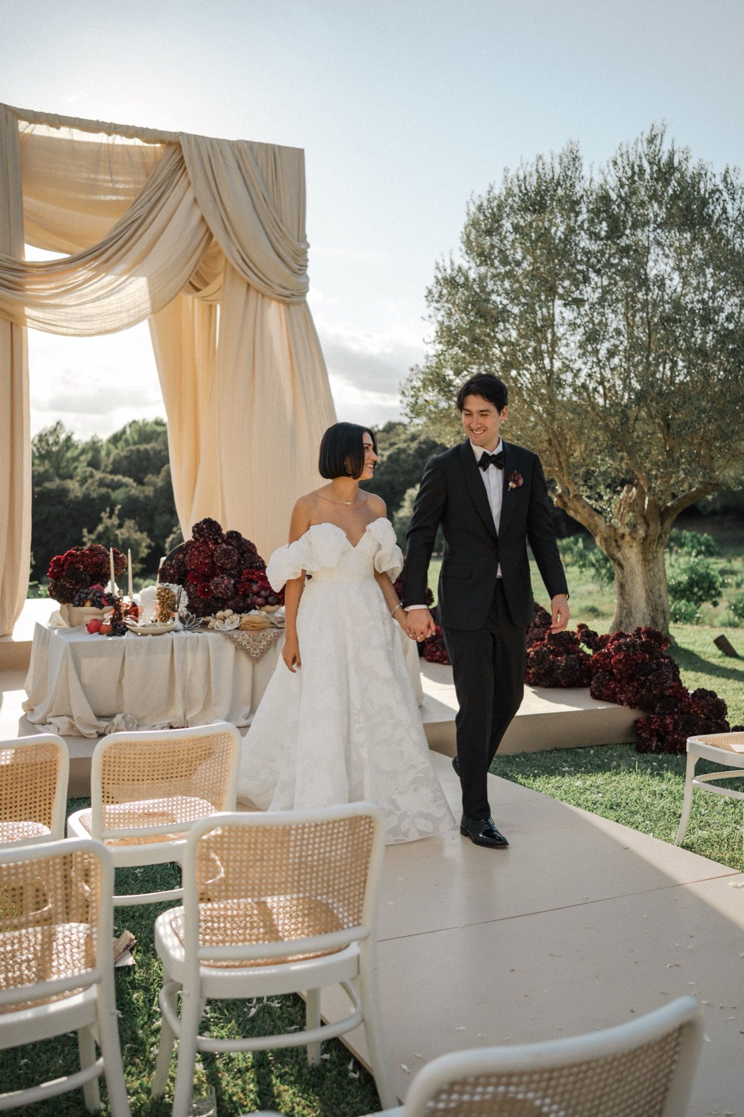 A romantic wedding scene in an elegant white draped pavilion with wicker chairs and olive trees in the background.