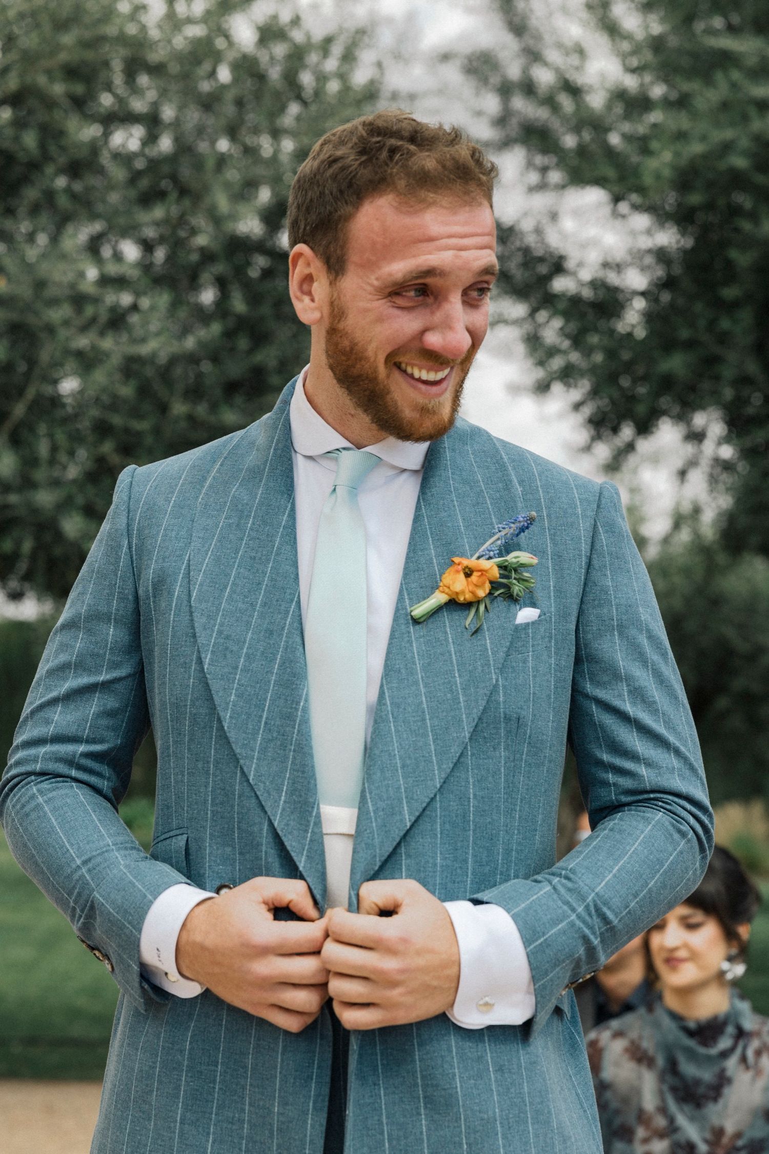 A groom in a light blue pinstripe suit and white shirt smiles while adjusting his jacket during an outdoor wedding.