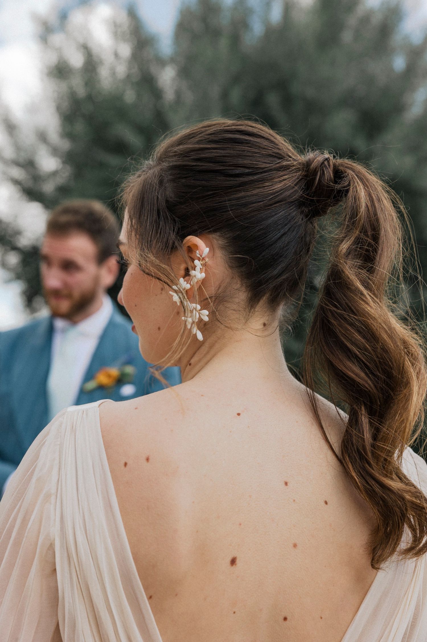 Back view of bride wearing delicate floral ear cuff and ponytail hairstyle at outdoor wedding ceremony.