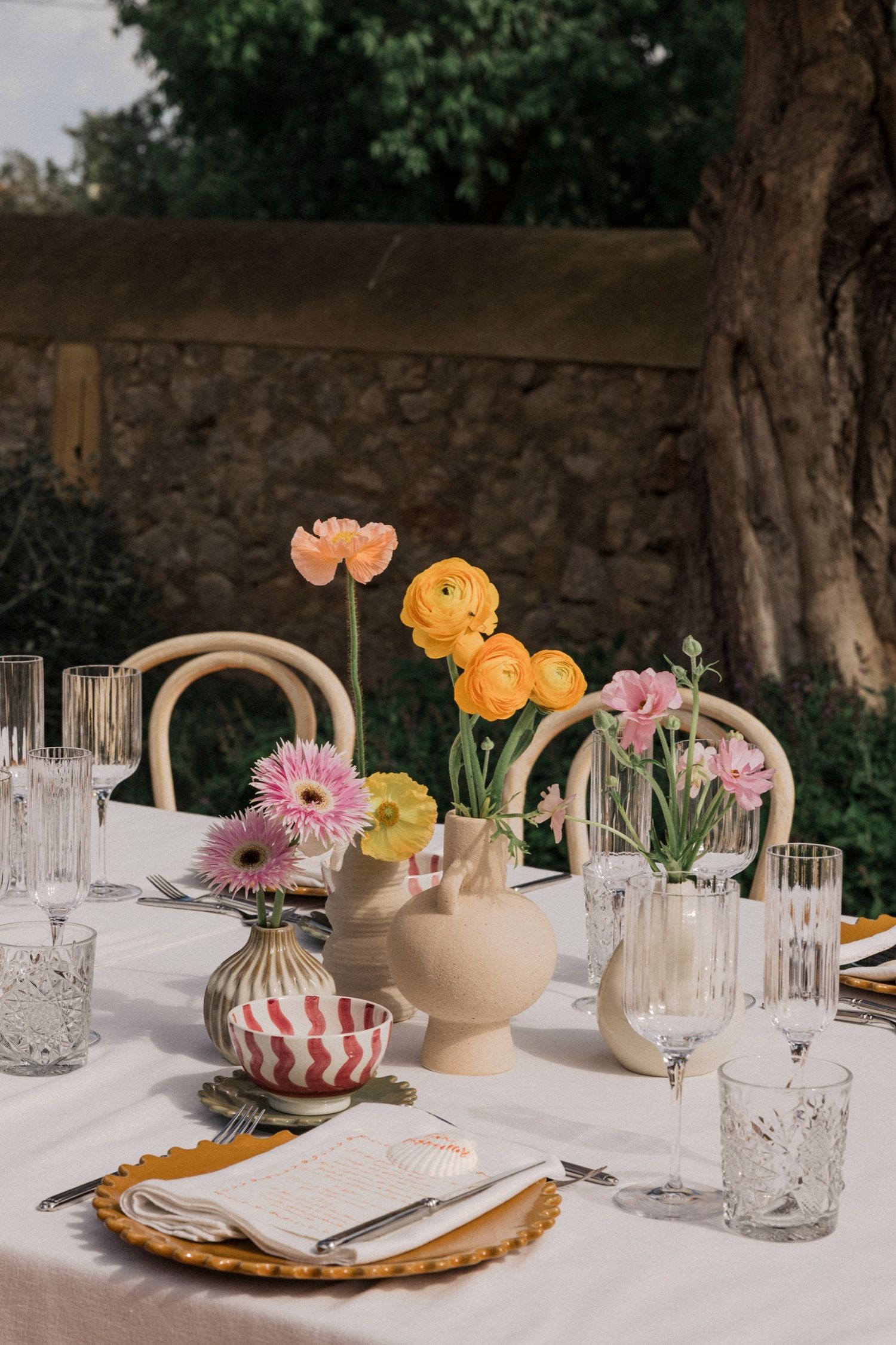 Elegant table setting with crystal glasses, gold chargers, and pink and orange flowers in delicate vases on white linens.