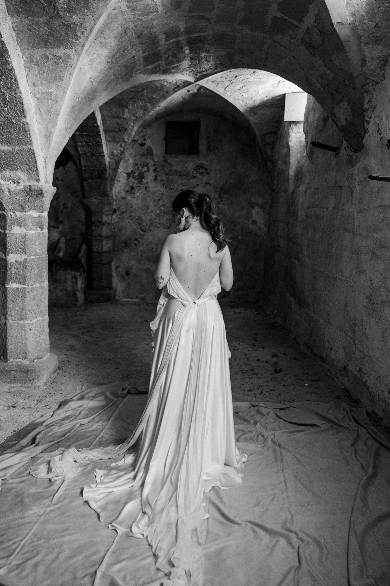 A black and white series showing a person in a white dress standing in an arched stone cellar.