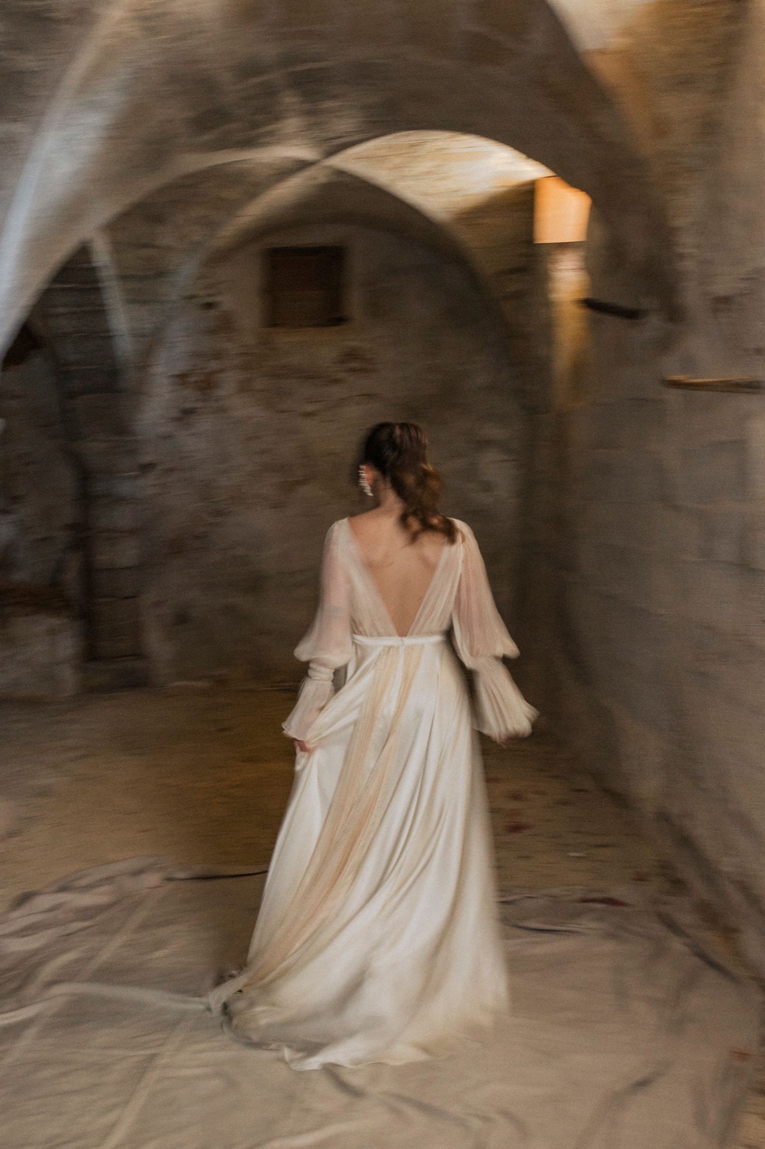 A figure in a flowing white dress moves ethereally through a stone archway in a dimly lit underground space.