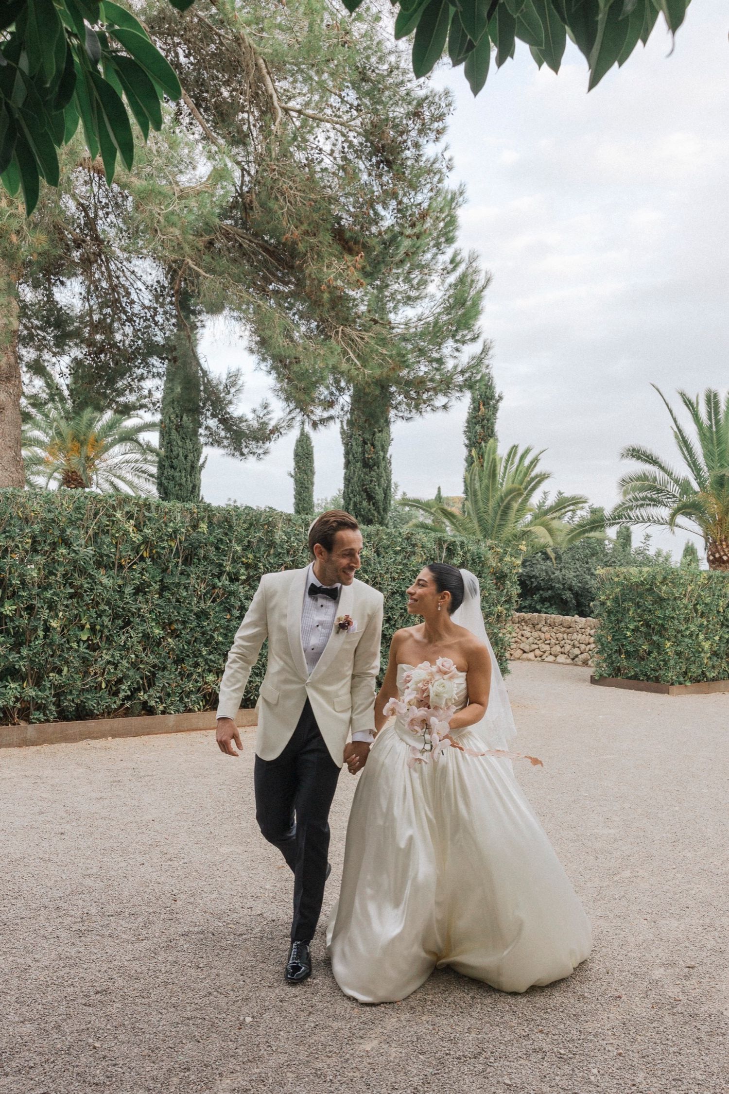 A bride and groom share romantic moments on a gravel path surrounded by lush greenery and palm trees.
