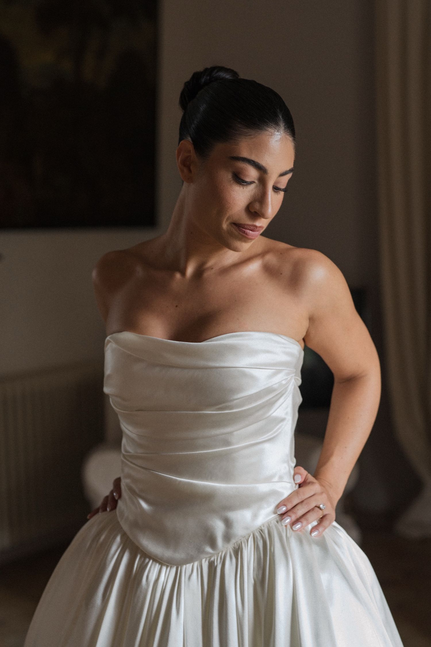A bride in a white strapless wedding gown with ruched bodice looks down thoughtfully in soft indoor lighting.