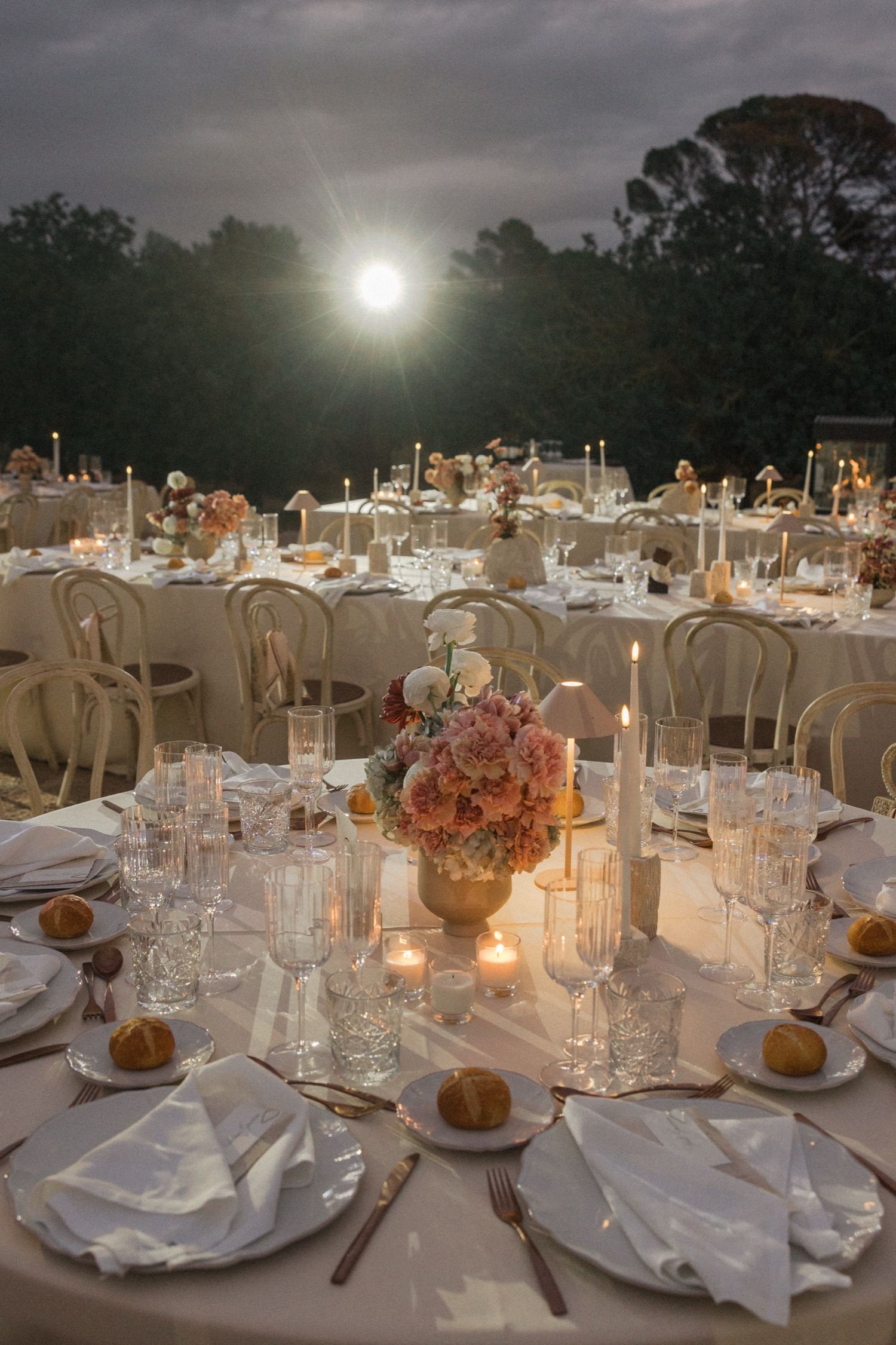 Elegant outdoor wedding reception tables set with white linens, floral centerpieces and glowing candles under evening sky.