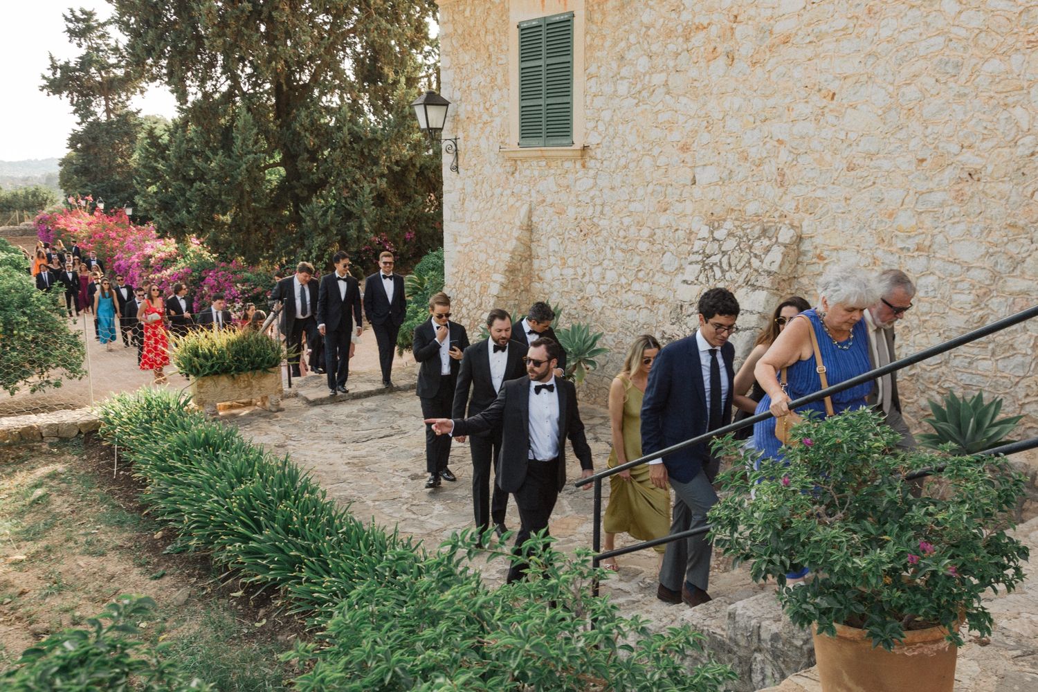 Wedding party and guests walking along a stone path lined with pink flowers and greenery at an outdoor venue.