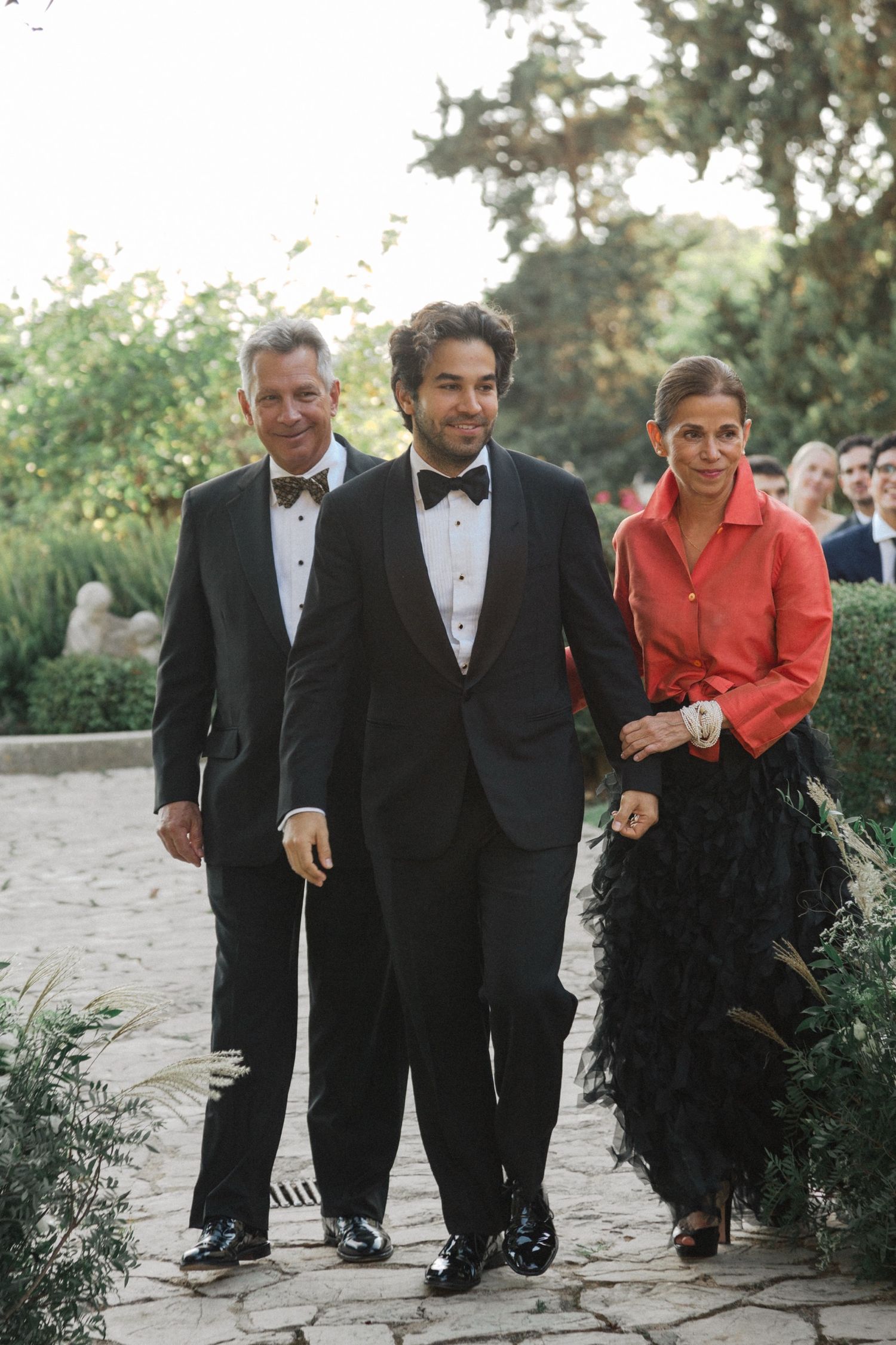 Three people in formal attire walking along a stone path at an outdoor event.