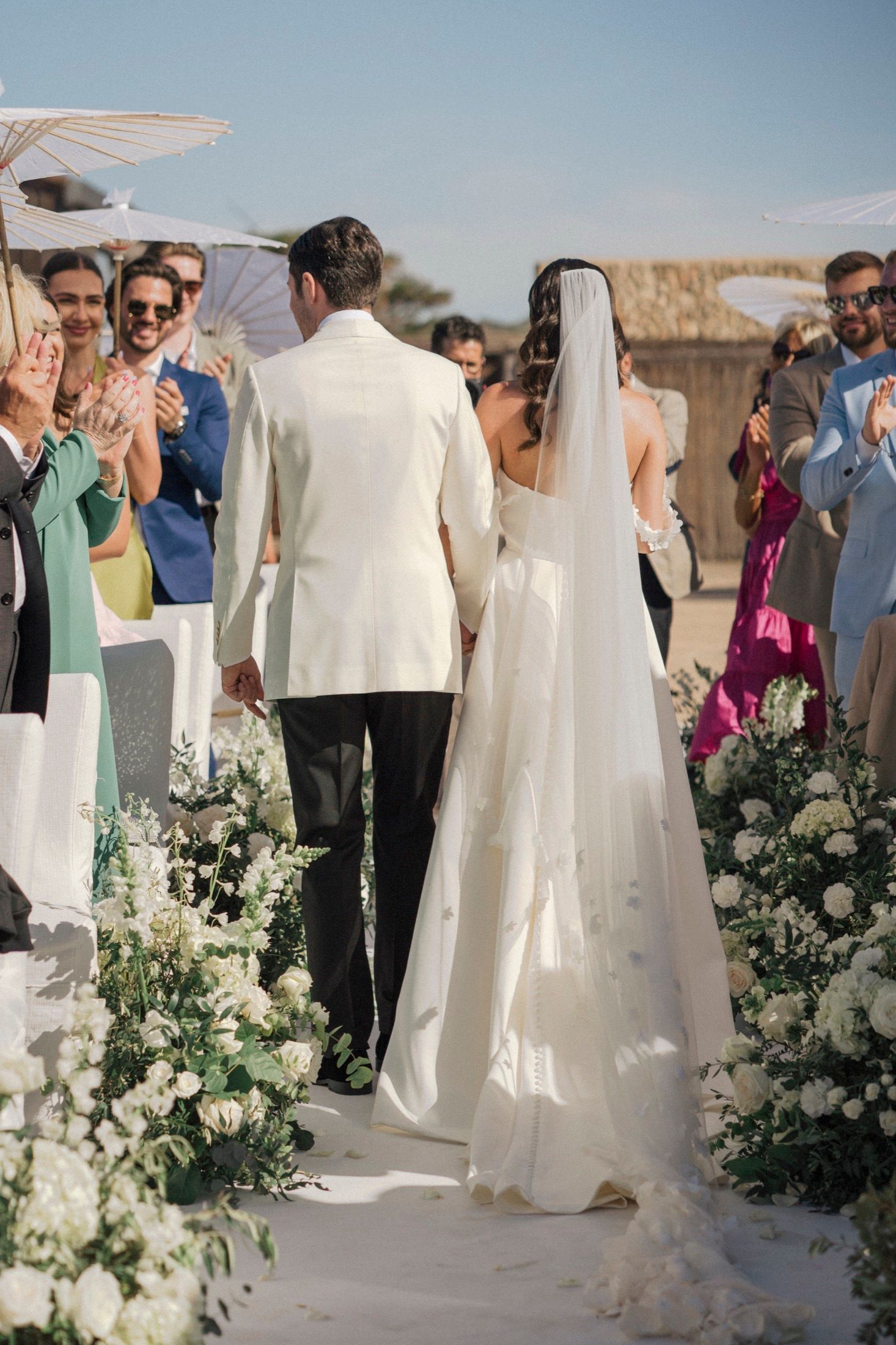 Bride and groom walk down outdoor aisle lined with white flowers, guests applauding at beach wedding ceremony.