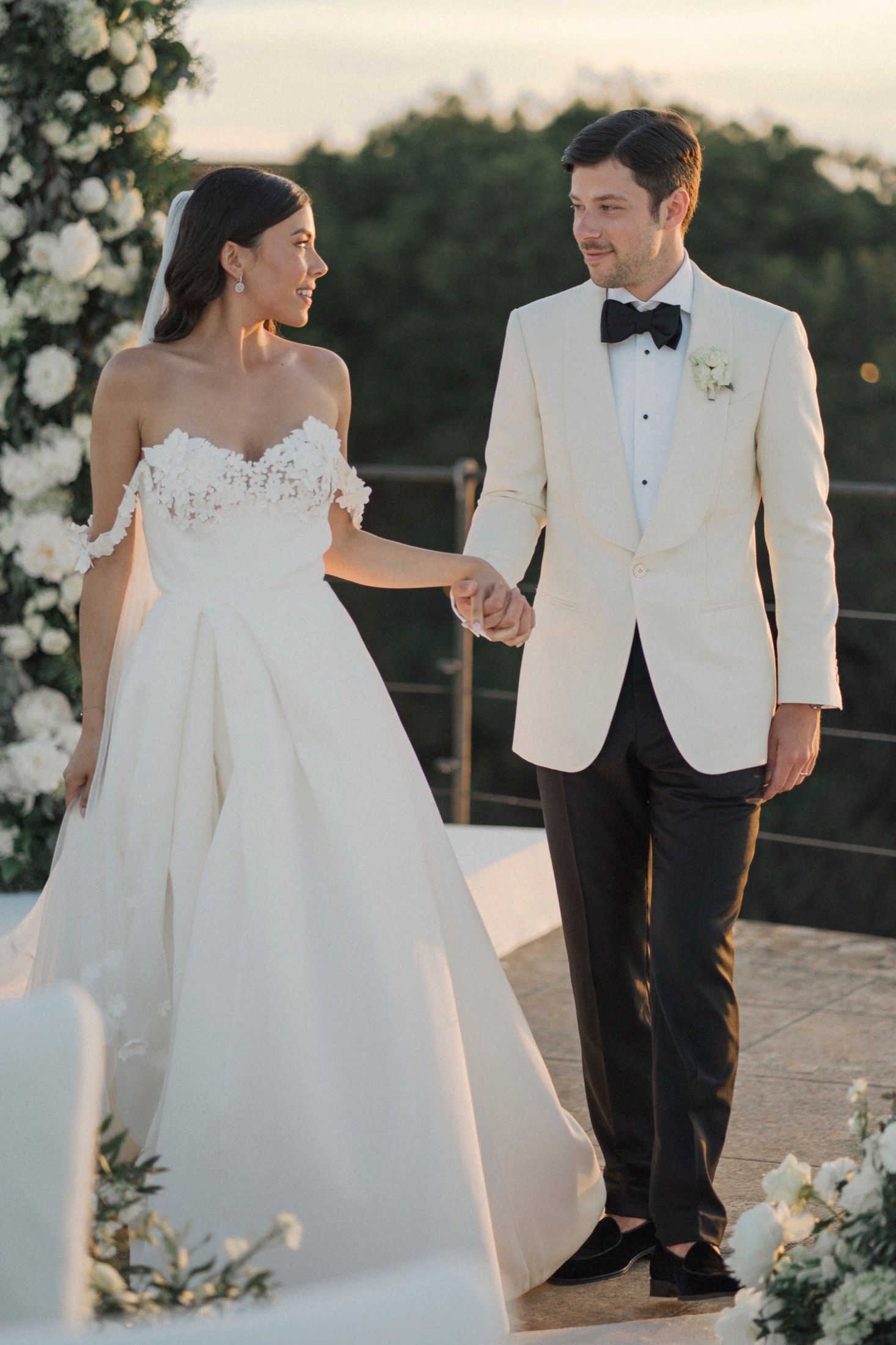 Bride in off-shoulder white gown holds hands with groom in ivory tuxedo at outdoor wedding ceremony.