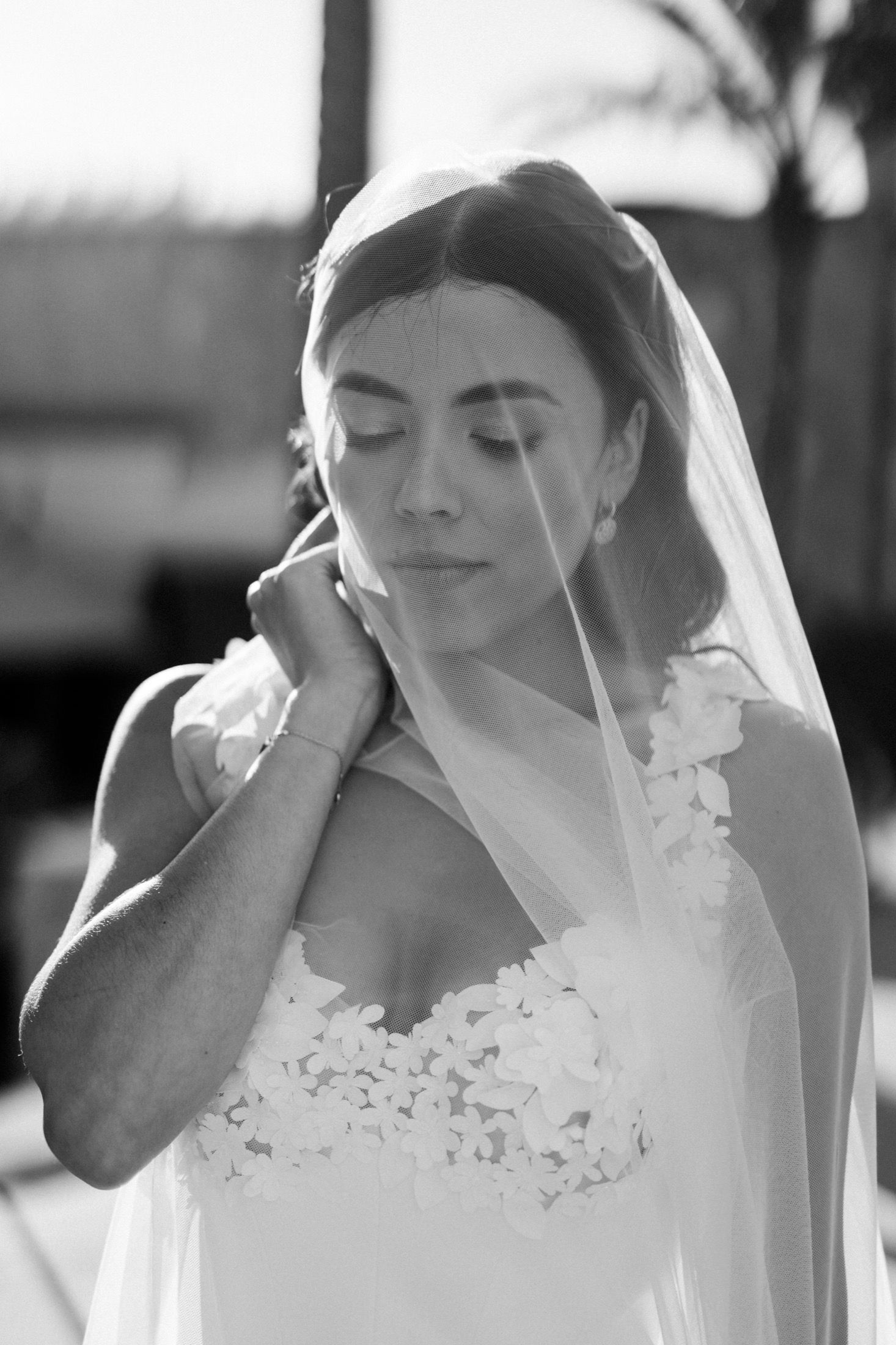 Black and white portrait of a bride in lace gown adjusting her veil with eyes closed.