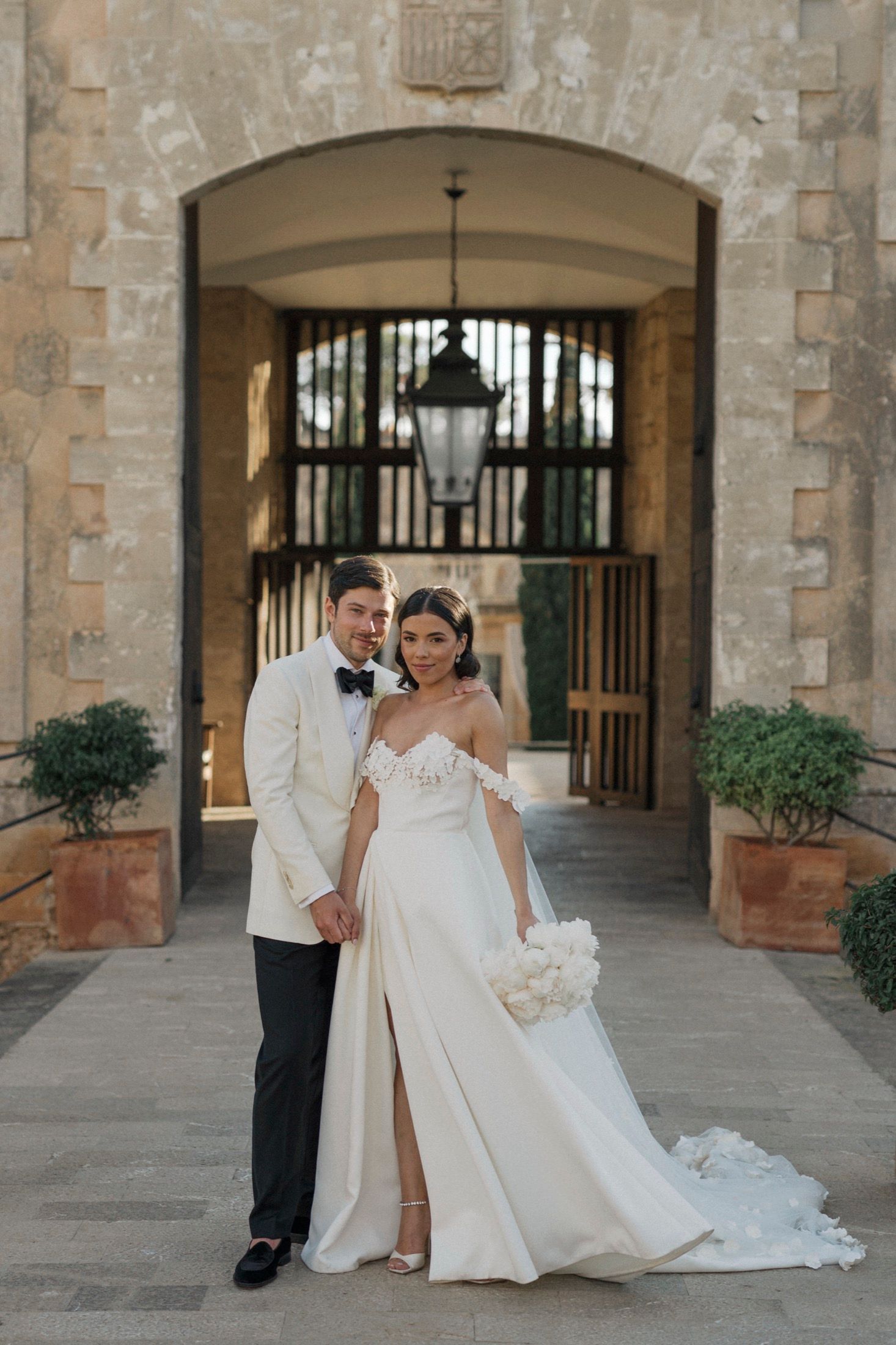 Bride and groom pose elegantly before a stone archway, she in an off-shoulder gown holding white blooms.