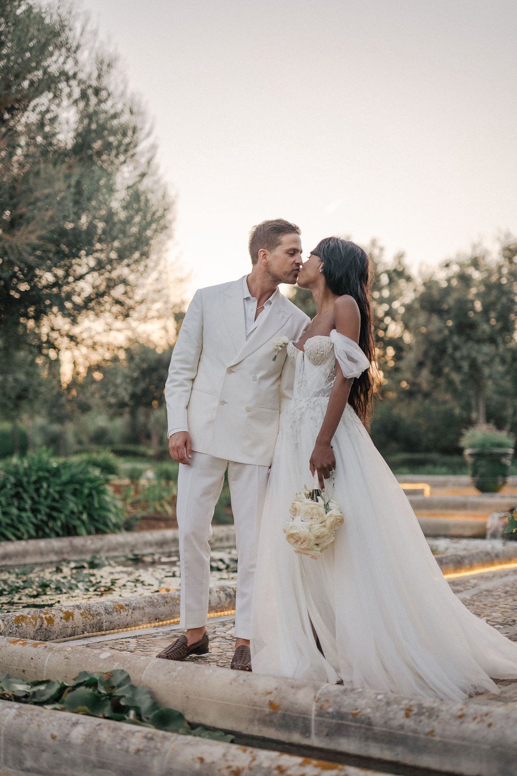 Elegant wedding couple in white attire posing by stone wall at sunset.