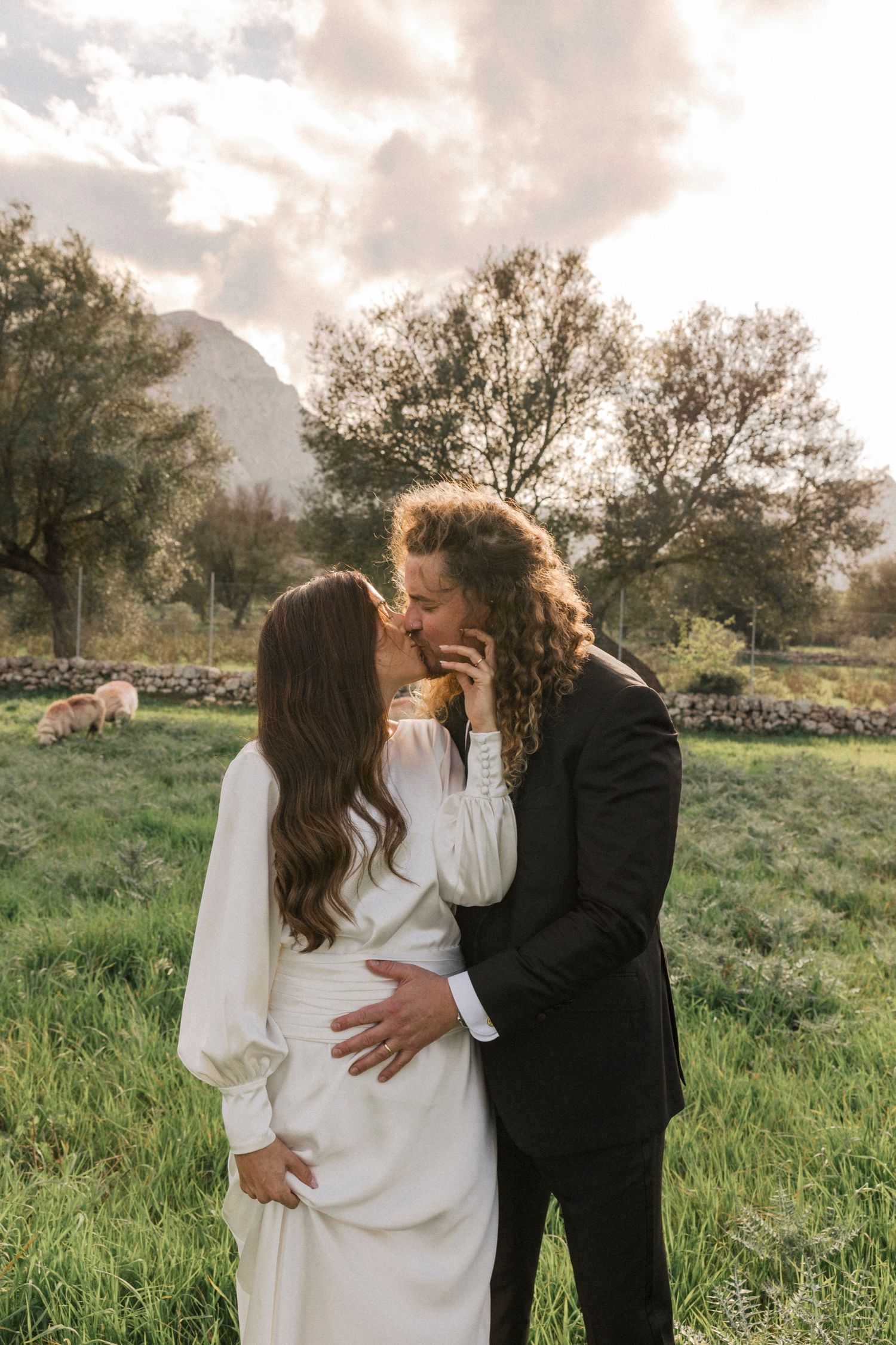 Couple embraces in a serene meadow with mountains and sheep in the background.