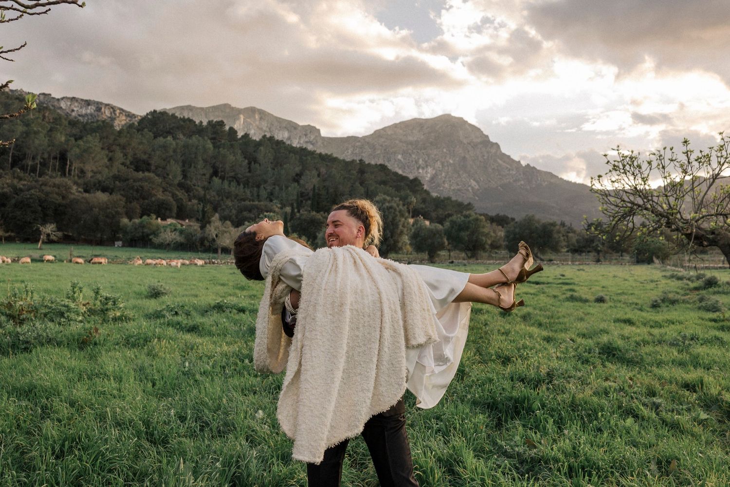 Newlyweds dance in a green field with mountains in the background during sunset.