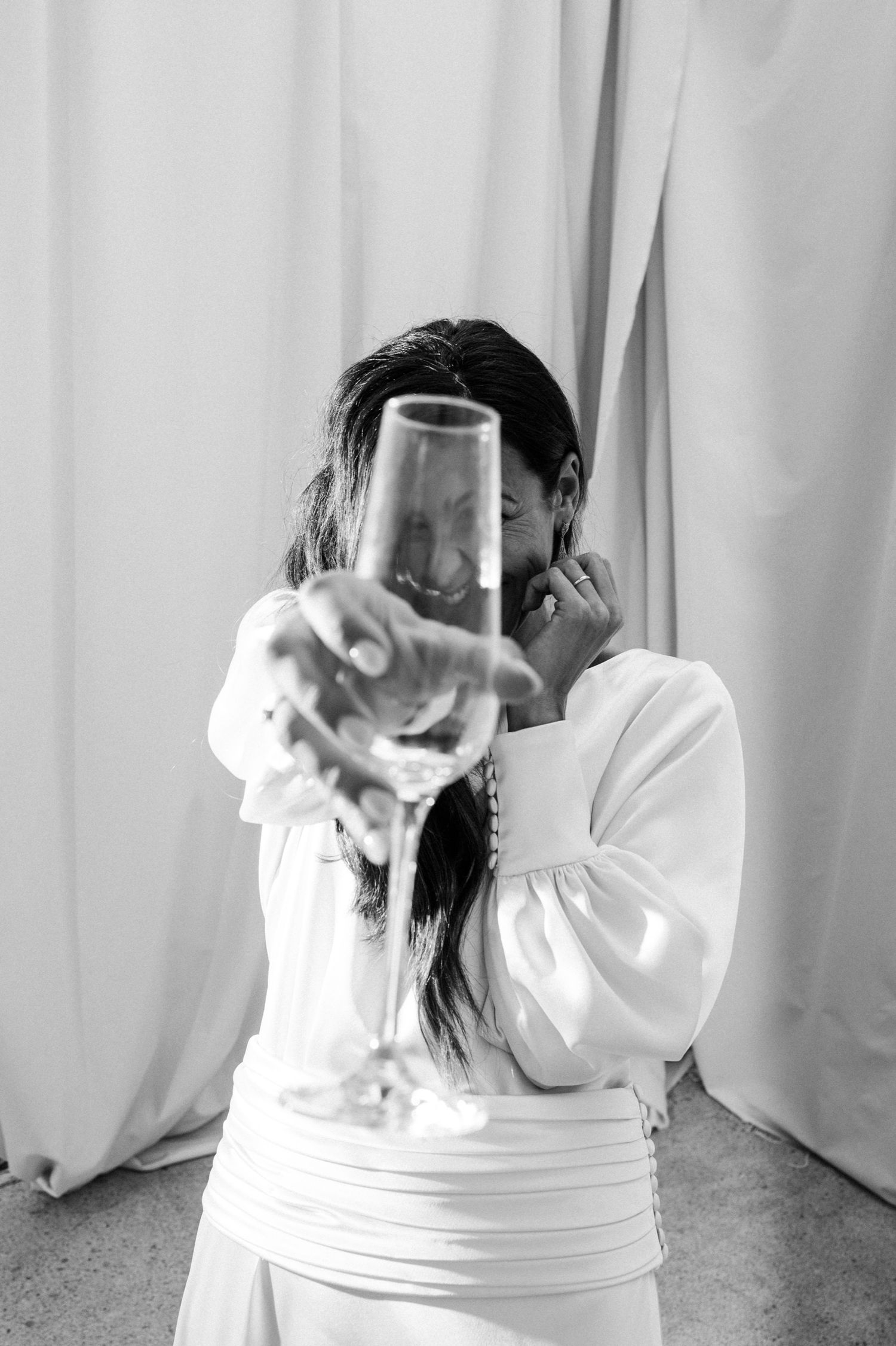 A person in a white dress holds up a champagne glass against a light backdrop.