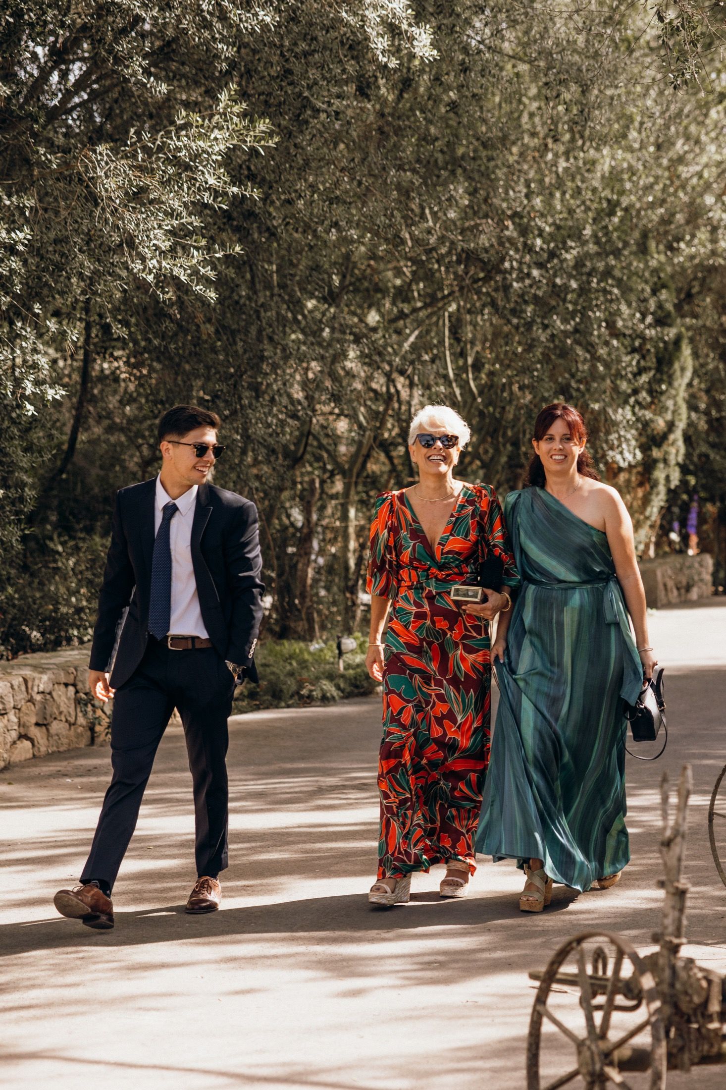 Three wedding guests in formal attire walk together on a sunlit outdoor path, wearing a suit, floral dress and green gown.