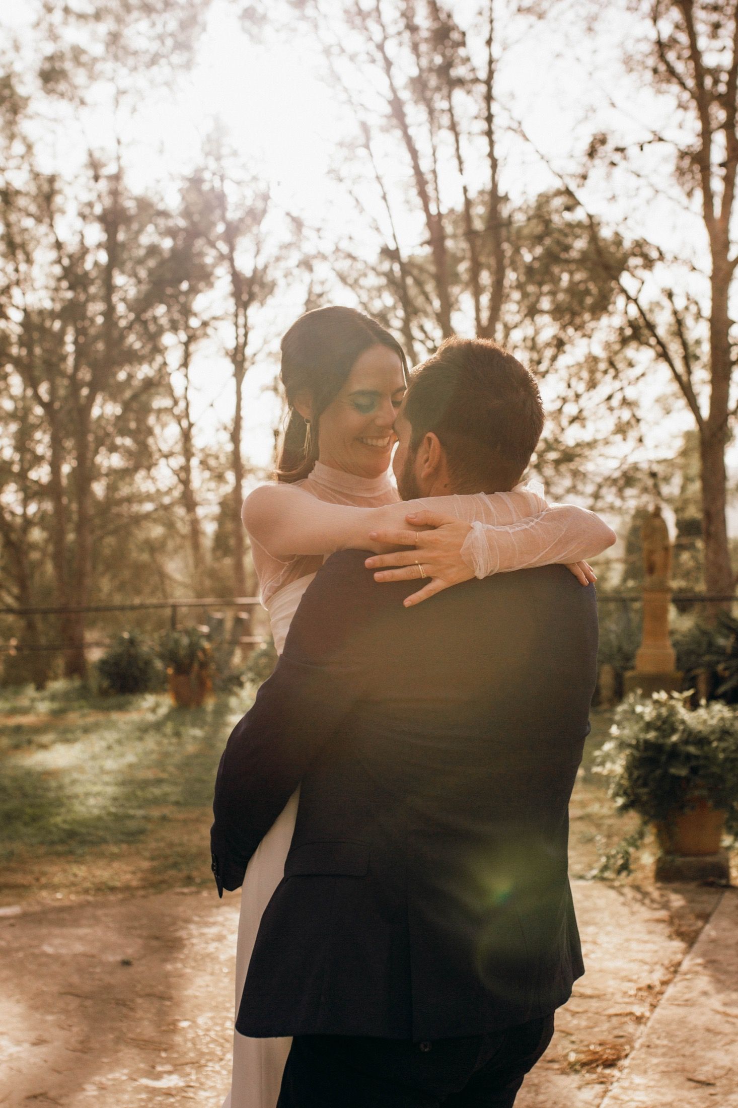 A romantic couple embraces outdoors during golden hour with sunlight filtering through trees in a garden setting.