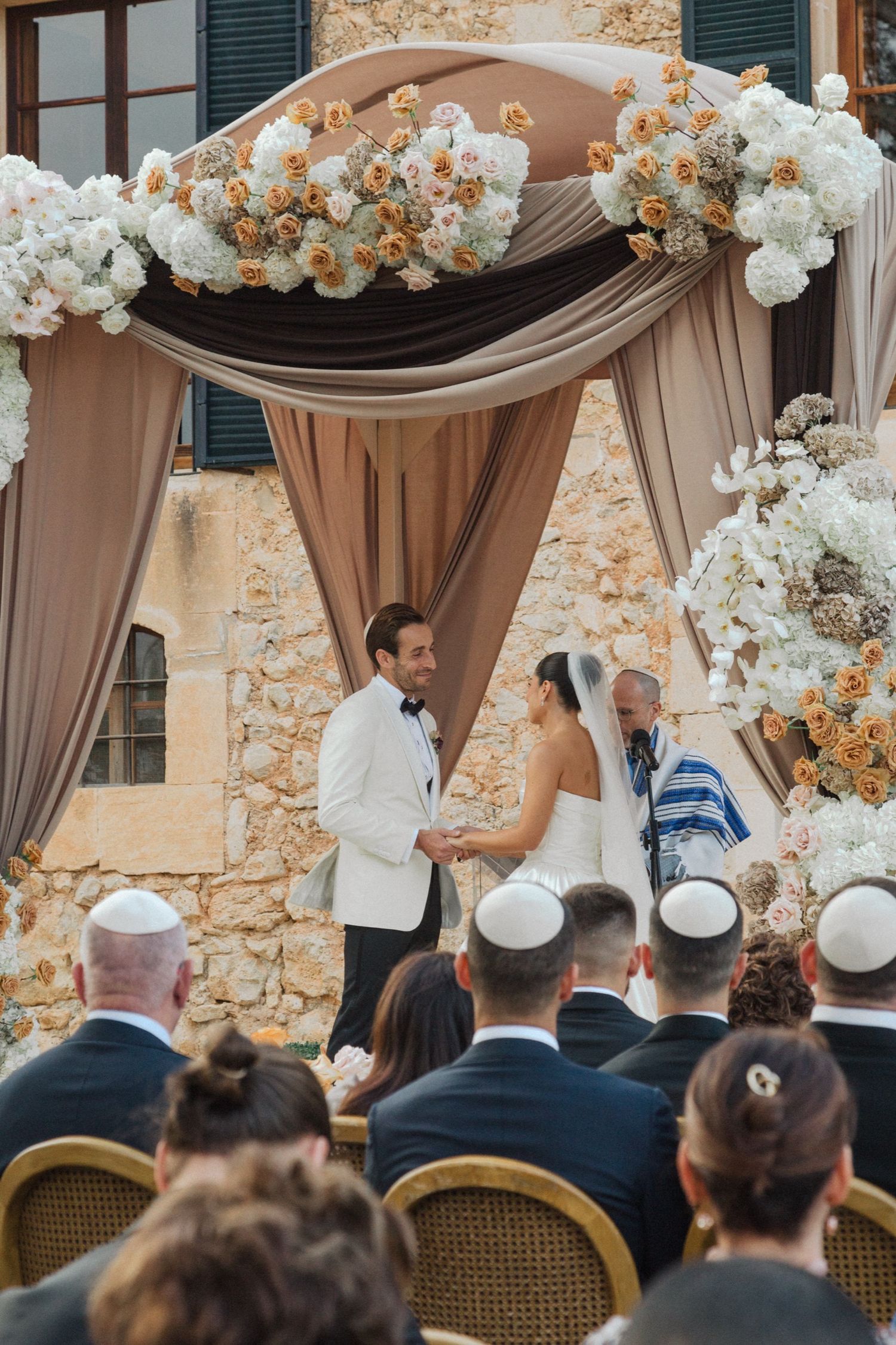 Elegant outdoor wedding ceremony under draped beige fabric arch adorned with white and peach floral arrangements.