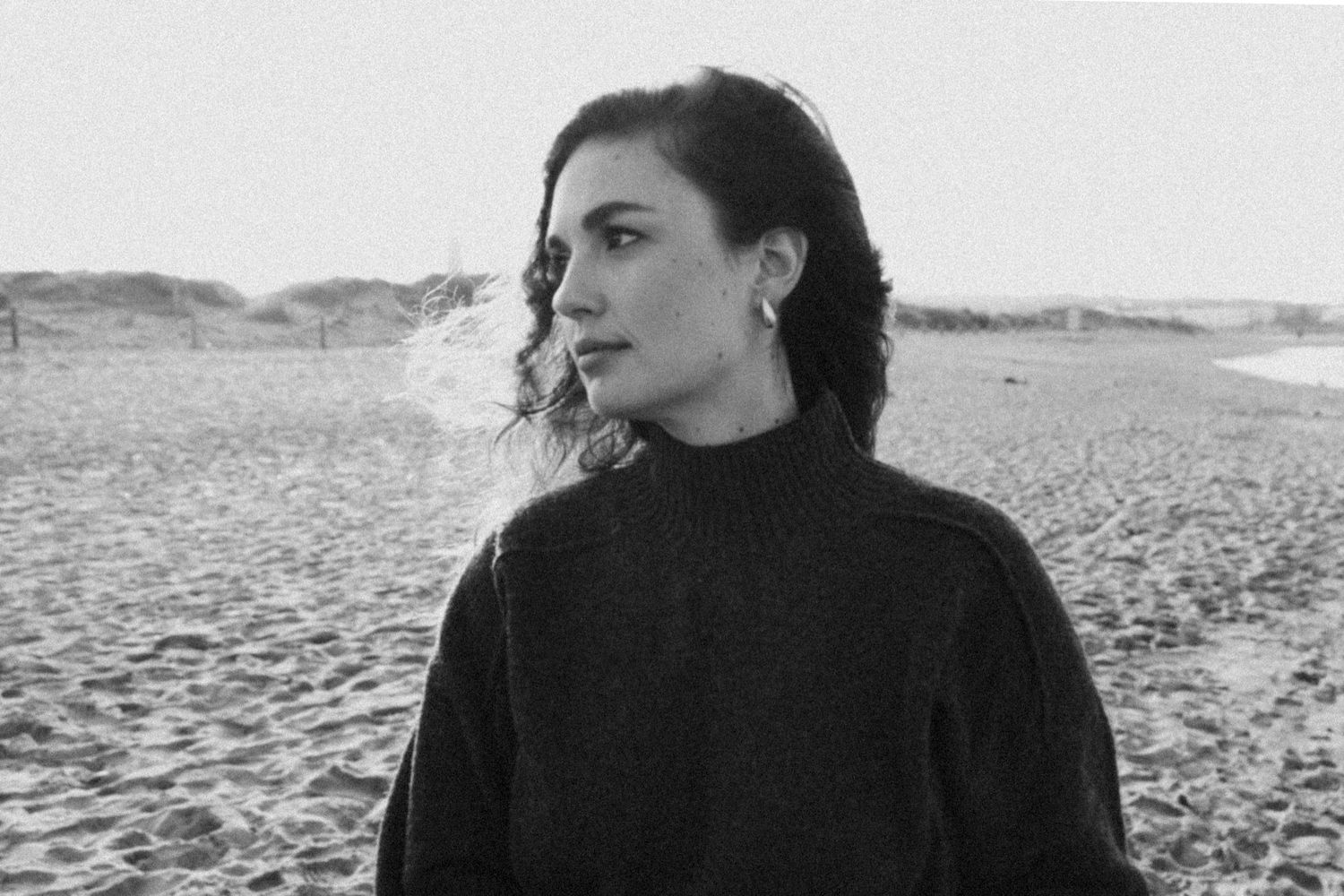 Black and white photo of someone in a turtleneck sweater looking to the side on a beach with dunes in background.