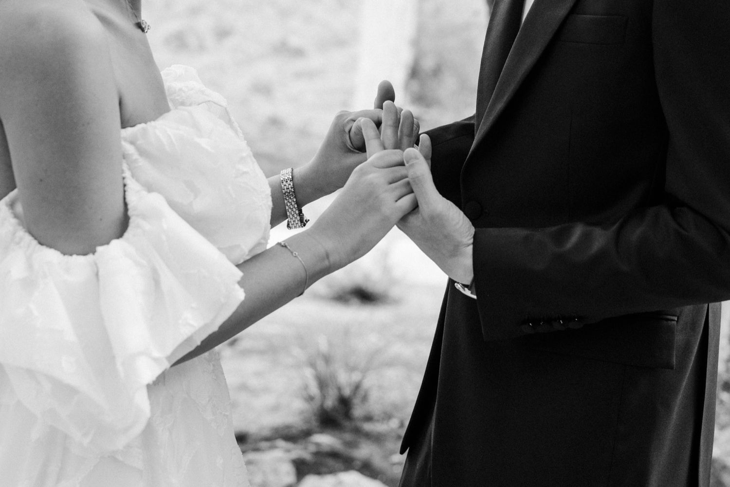 Black and white closeup of hands exchanging wedding rings during an outdoor ceremony.