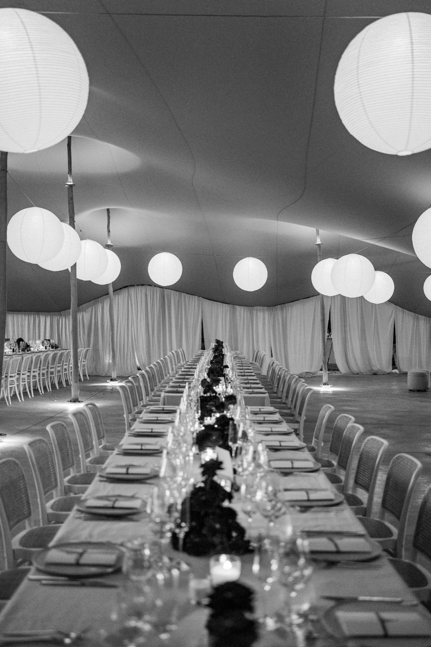 Long elegant banquet tables set with white linens and paper lanterns hanging from above in a modern tent reception space.