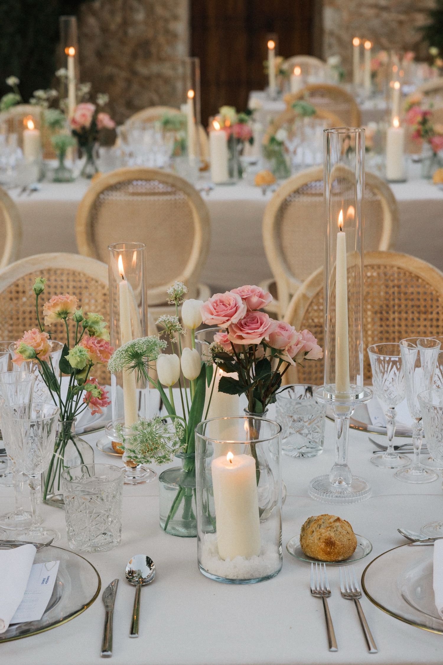 Elegant wedding table settings with pink roses in glass vases, white candles and rattan chairs create a romantic atmosphere.