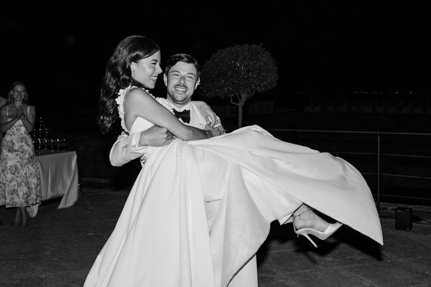 Groom carrying bride in white wedding gown outdoors at night in black and white photo.