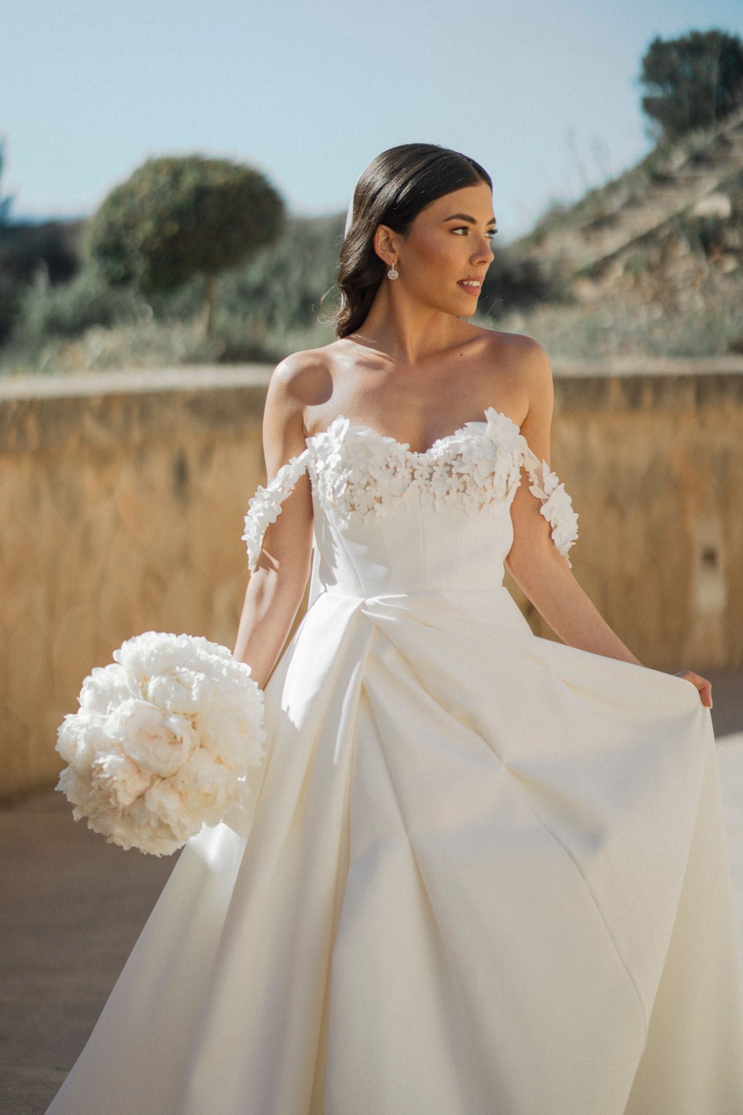 Bride in off-shoulder white lace ball gown holding white peony bouquet outdoors in sunny Mediterranean setting.
