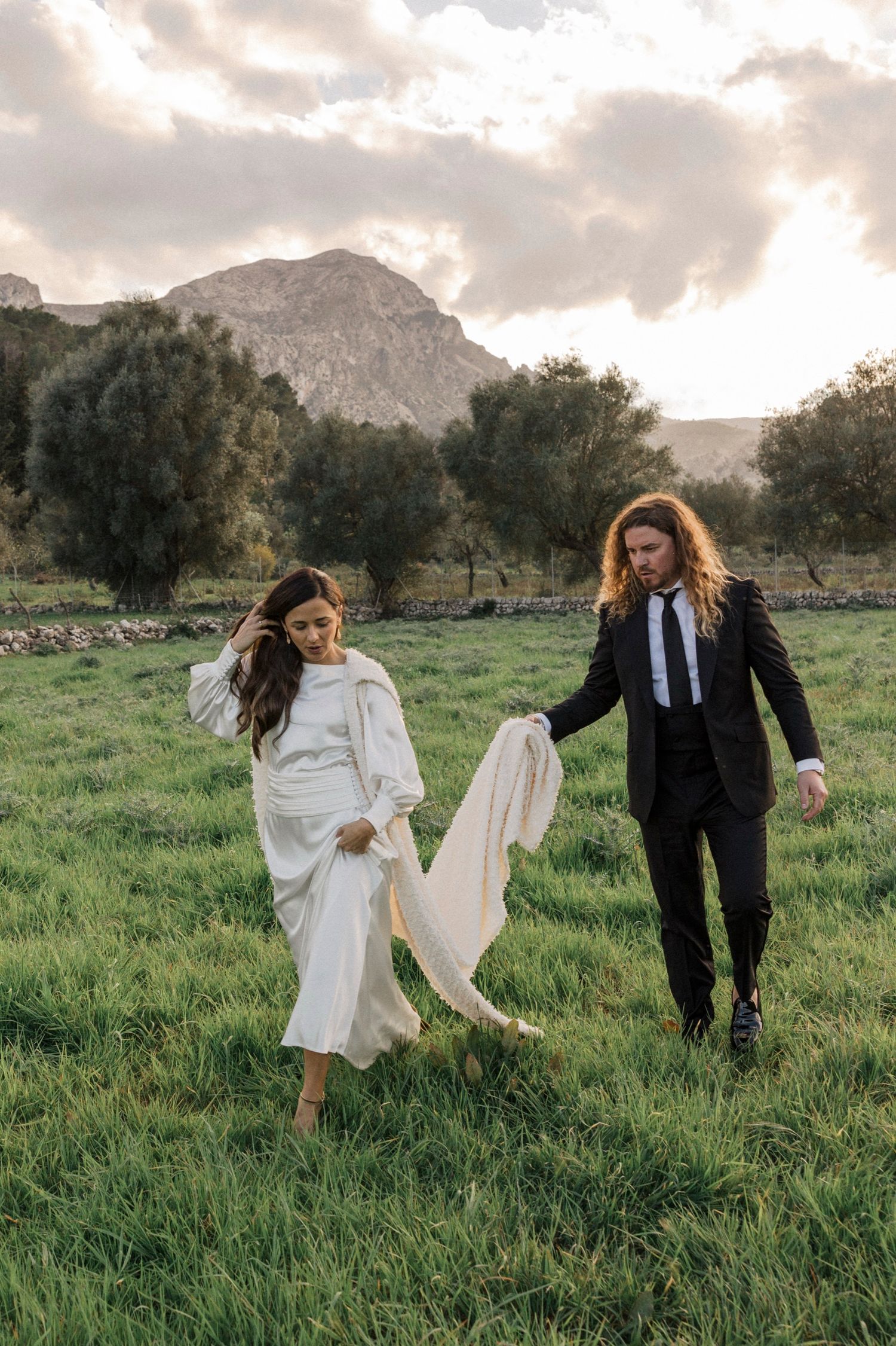 A couple playfully walks through a green field with mountains and dramatic clouds in the background.