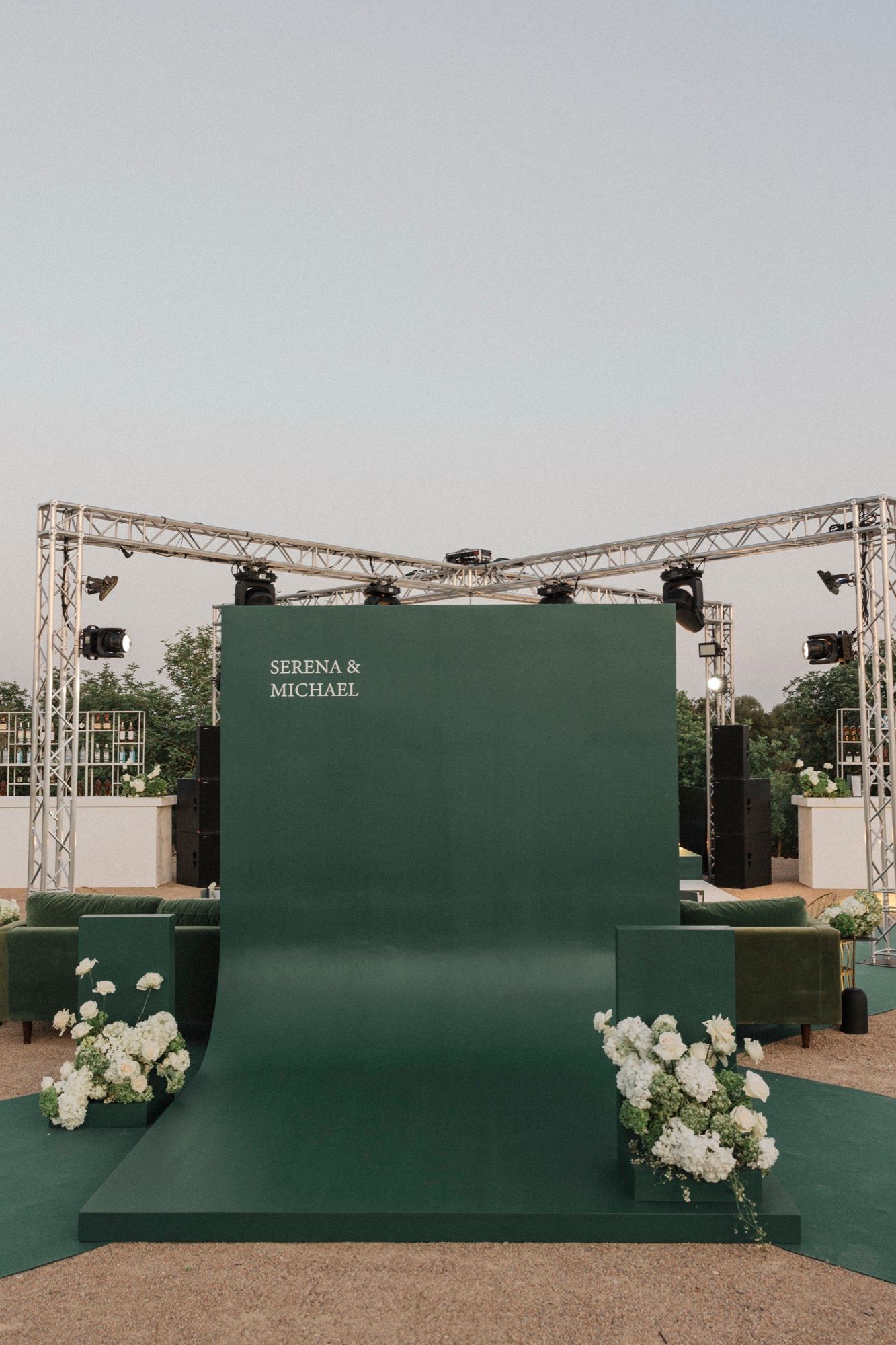 Modern dark green photo backdrop with white floral arrangements at outdoor wedding venue.