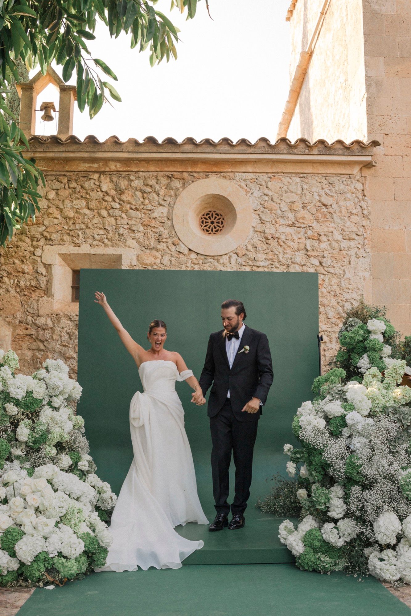 A wedding couple poses against a stone wall backdrop with elegant white floral arrangements and a green backdrop.