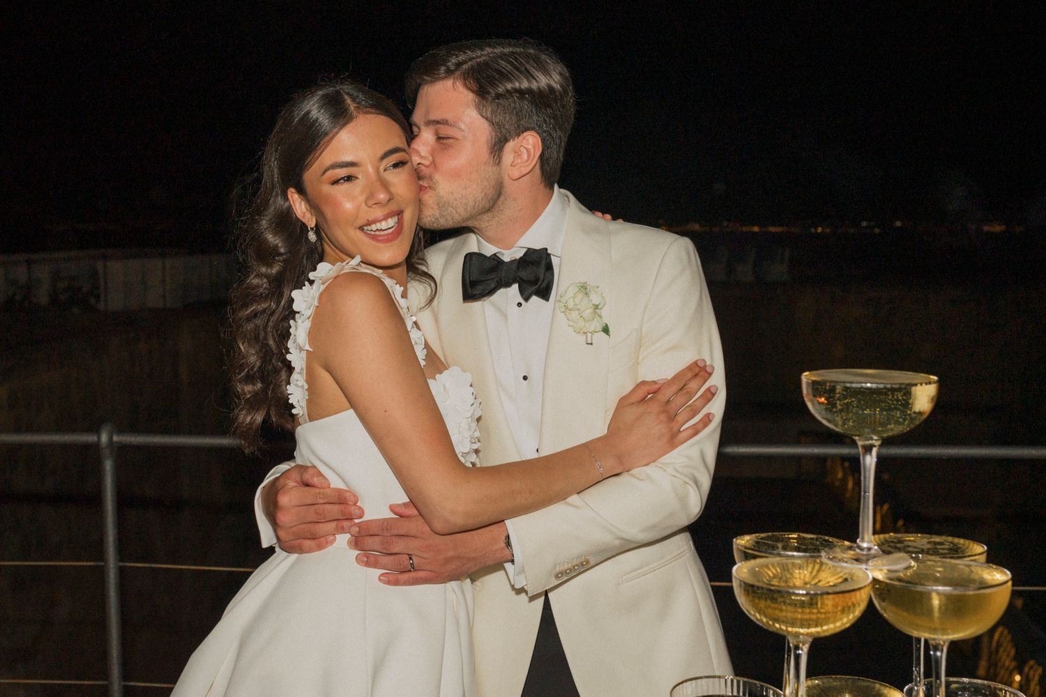 Newlywed couple in white wedding attire sharing a kiss beside a champagne tower at a romantic evening reception.