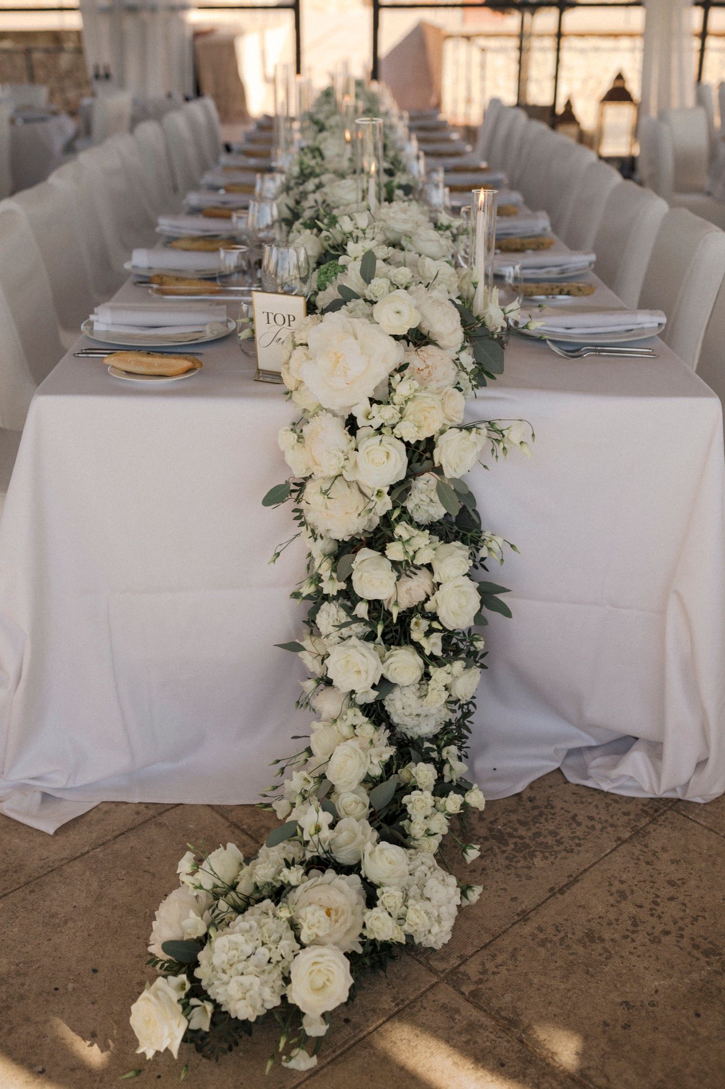 Elegant wedding reception table with lush white floral garland runner, white roses, greenery, gold chargers, and neatly folded napkins.