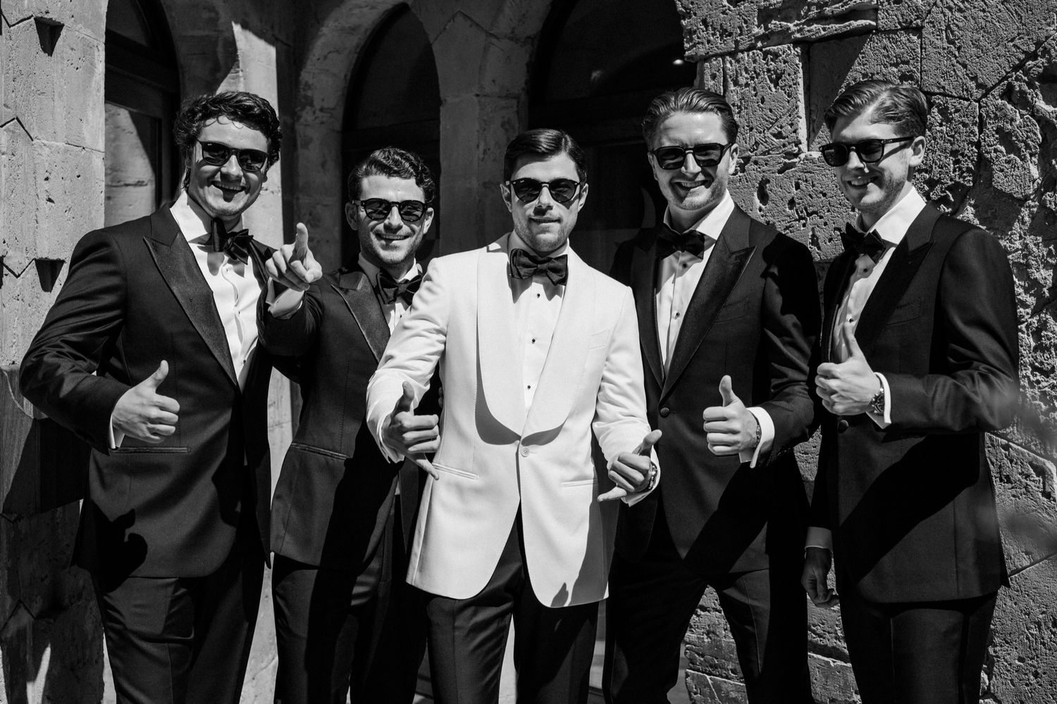 Five men in tuxedos and sunglasses pose playfully in black and white photo, likely a wedding party.