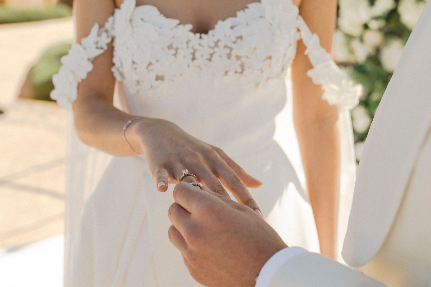 Groom placing a diamond ring on bride's finger during outdoor wedding ceremony.